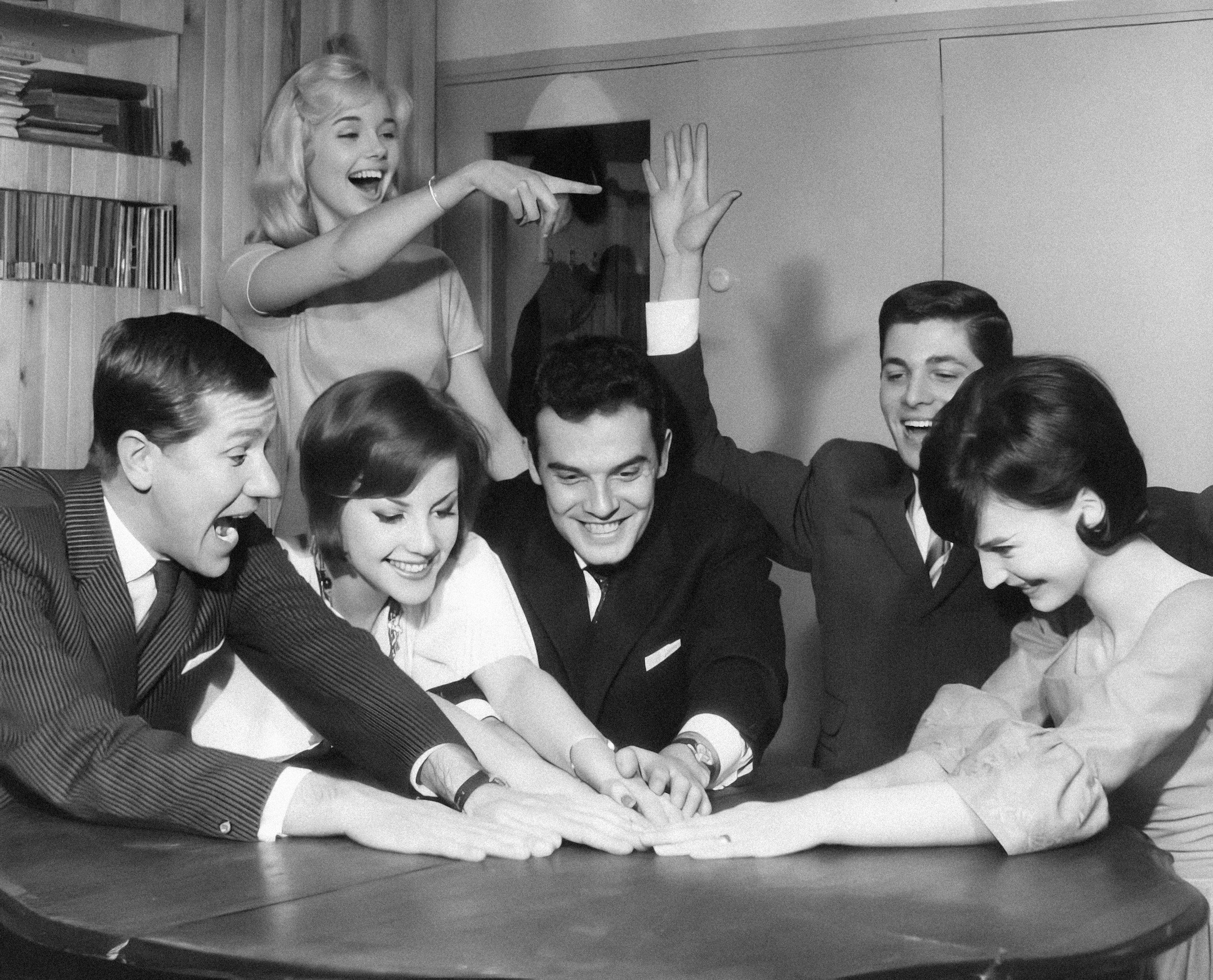 Un gruppo di persone sorridenti si tiene per mano e si abbraccia sopra un tavolo. È una scena di allegria e unità in un ambiente interno.