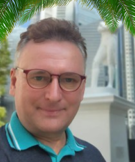 A man with glasses and short curly hair taking a selfie outdoors, with a background of modern buildings and green palm leaves.