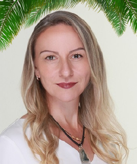 A woman with long blonde hair, wearing a white top and a necklace, posing in front of a backdrop with tropical palm leaves.