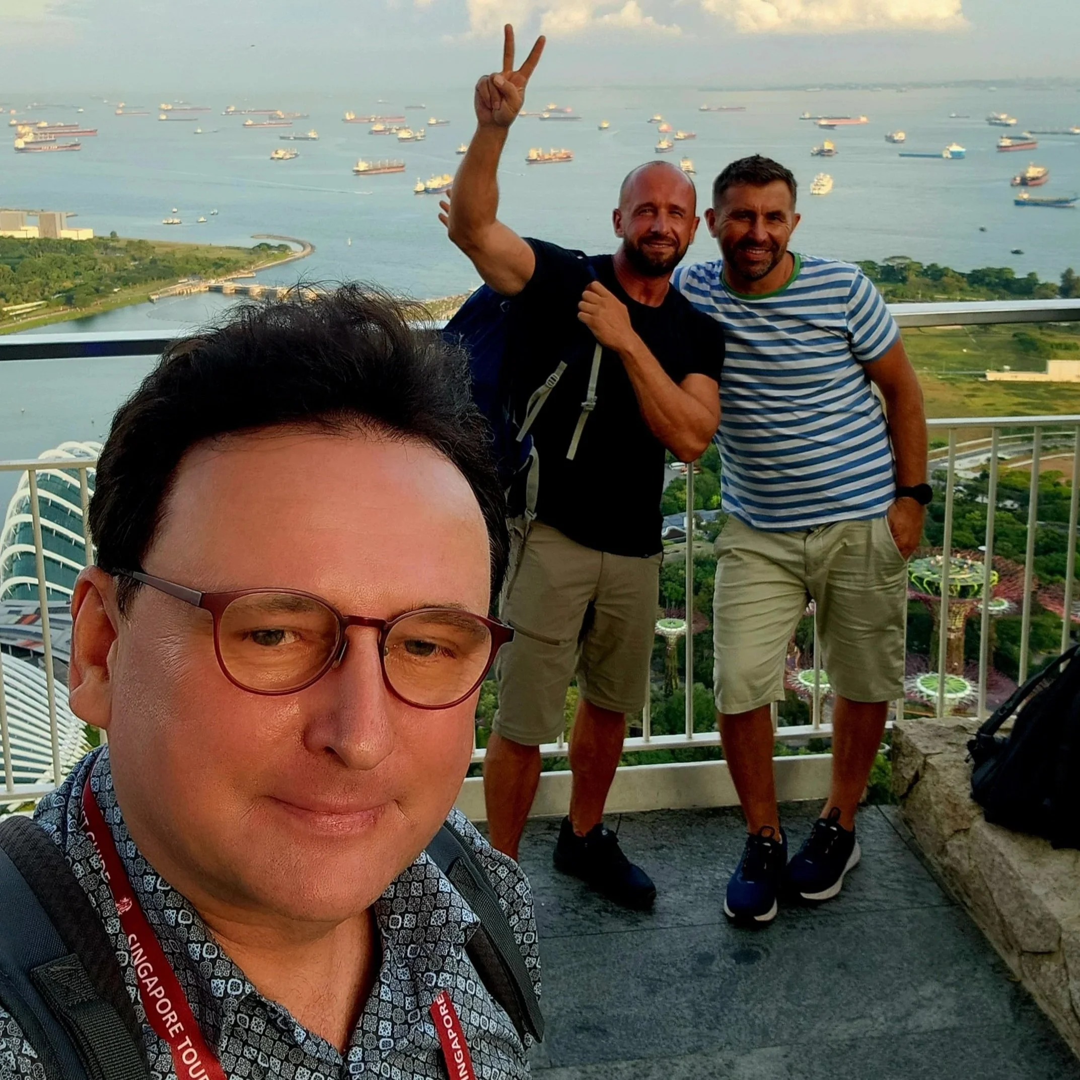 Two men and a woman standing outdoors on a balcony overlooking a body of water with numerous ships. One man is making a peace sign with his fingers, and the photo is taken during the daytime.