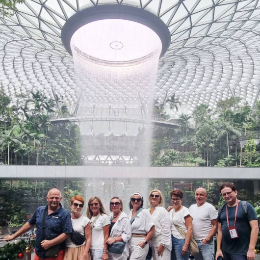 Group of ten people standing inside Jewel Changi Airport in Singapore, with a large indoor waterfall and lush greenery above them.