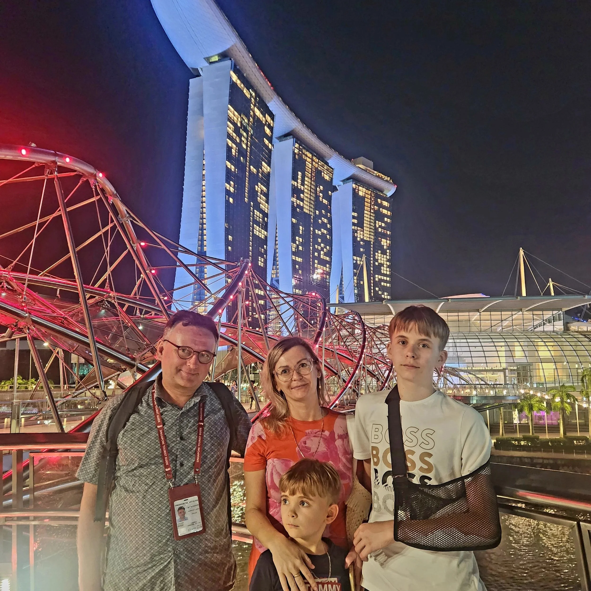 A group of five people, including children and adults, standing together in front of the Marina Bay Sands hotel and a Ferris wheel in Singapore at night, with city lights and high-rise buildings in the background.
