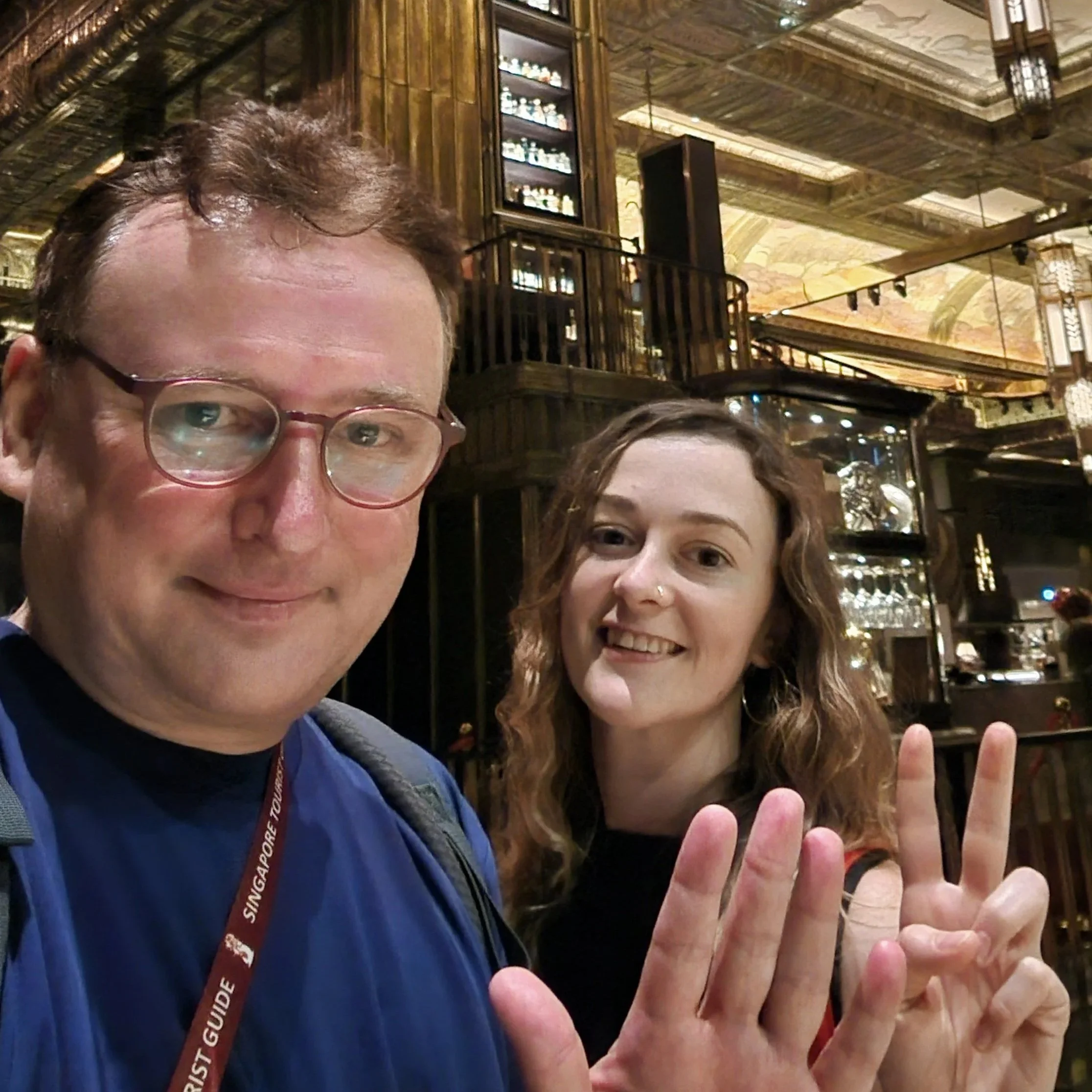Partial view of three people in a warm, ornate indoor setting with decorative wooden and gold accents; two individuals are smiling, one is giving a thumbs-up gesture.