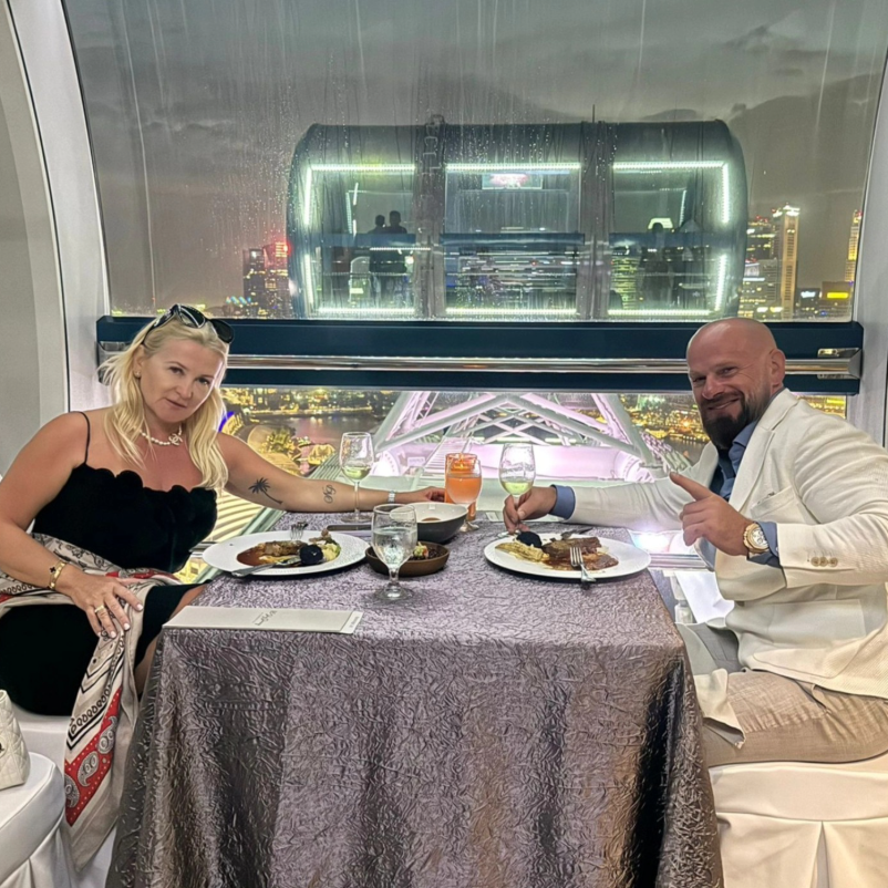 A man and woman dining at a restaurant with a city view at night. They are sitting at a table with plates of food and drinks, including wine and cocktails. The woman has blonde hair, sunglasses on her head, and wears a black top, while the man is bal