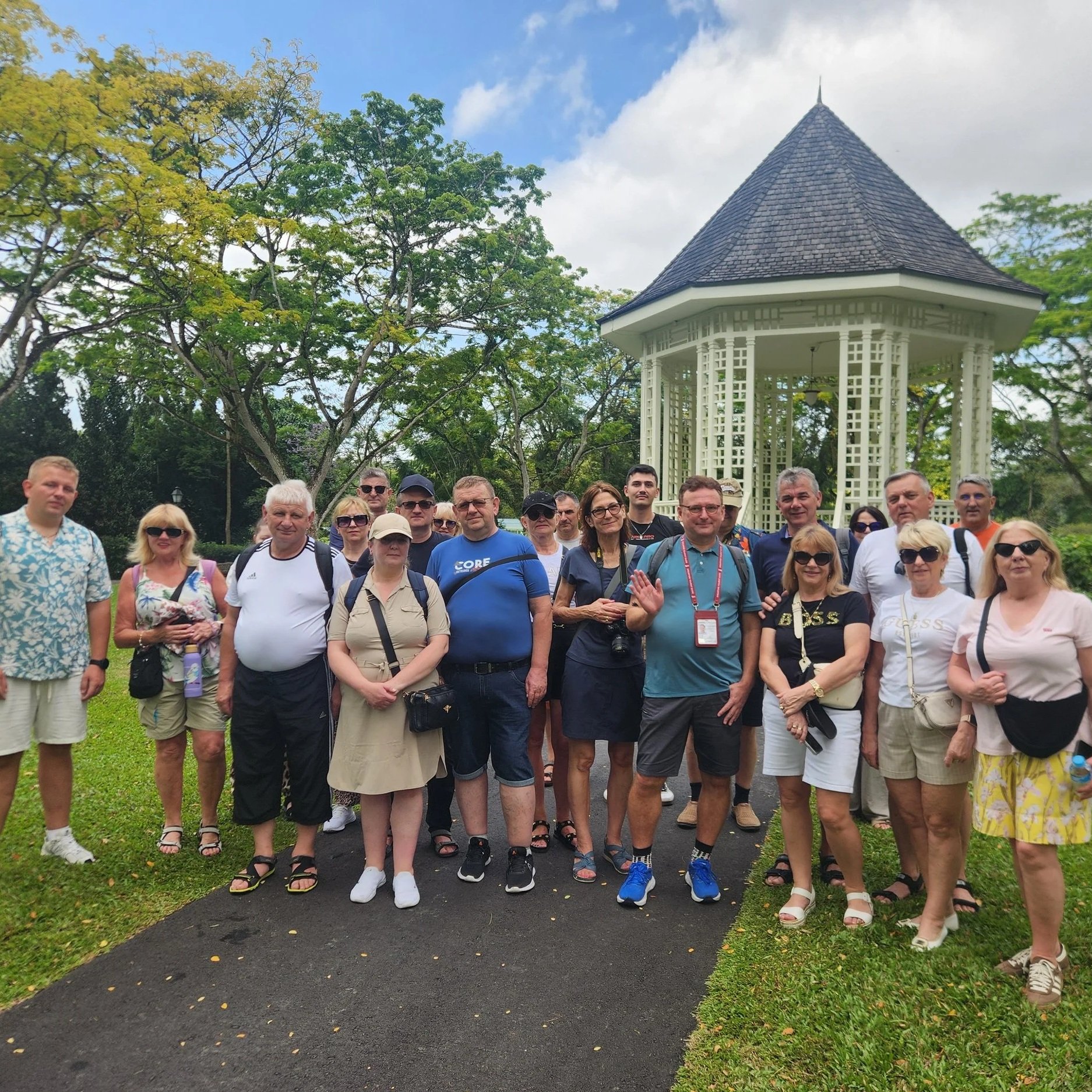 Group of diverse people standing outdoors in front of a gazebo, with trees and blue sky in the background.