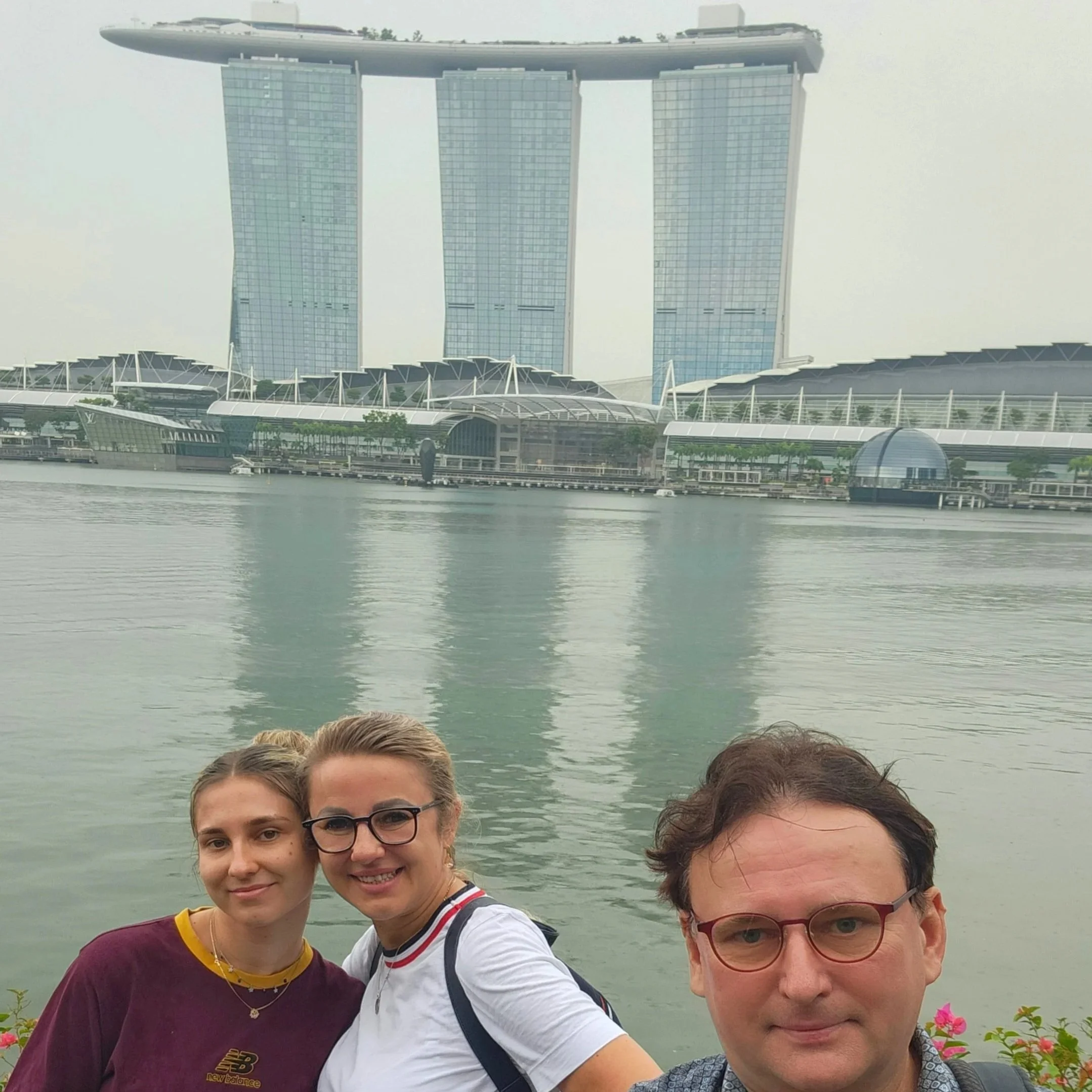 A group of people taking a selfie in front of Marina Bay Sands hotel in Singapore, with the water of Marina Bay and some plants in the foreground.