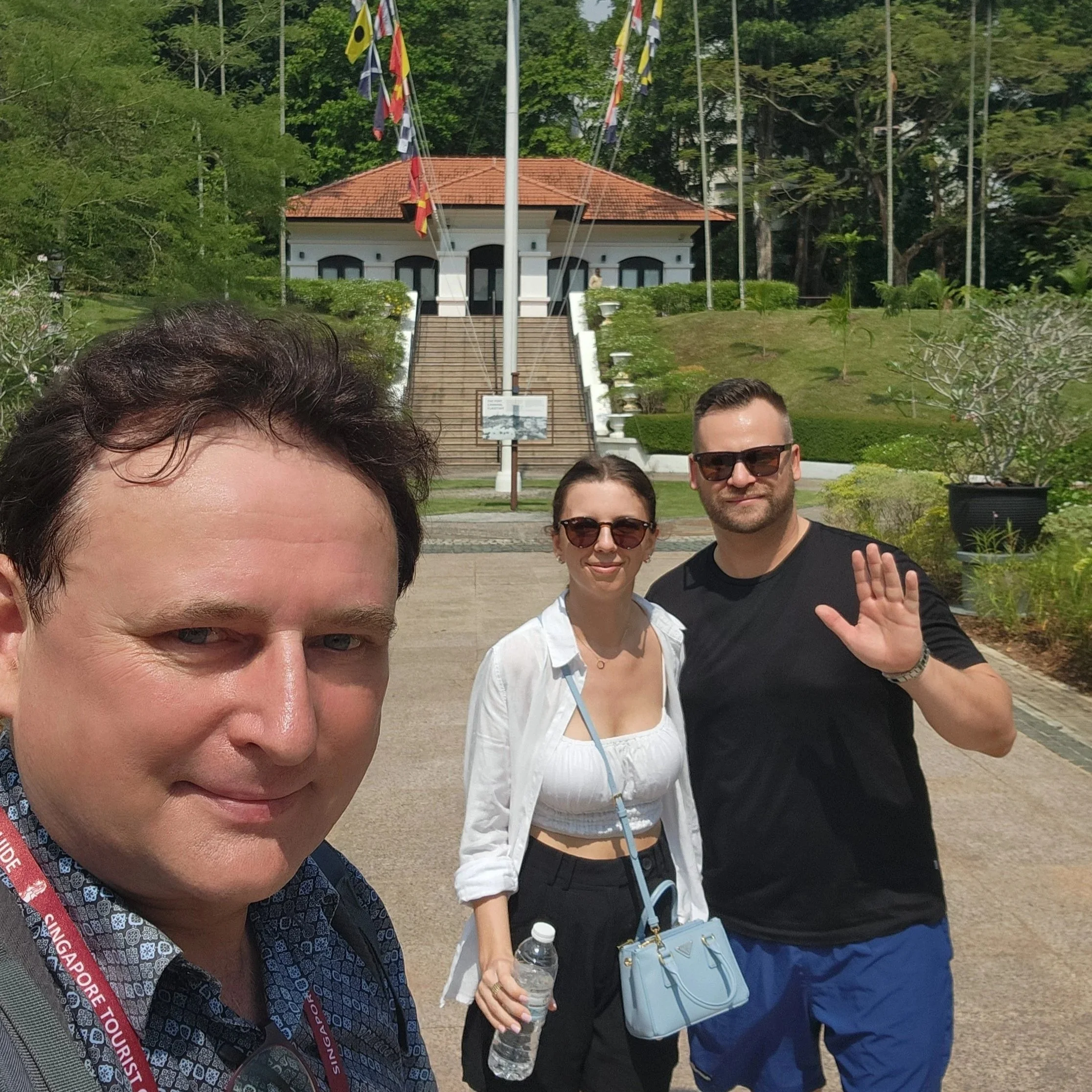 Group of people of diverse ages and genders posing outdoors in front of a staircase leading to a building with a red tile roof, with several flags flying on poles and surrounded by lush green trees.