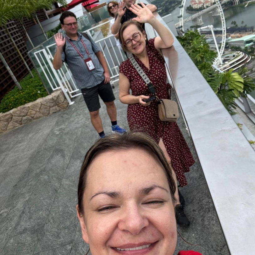 A group of five people taking a selfie on a rooftop with a Ferris wheel and river in the background, smiling and waving.