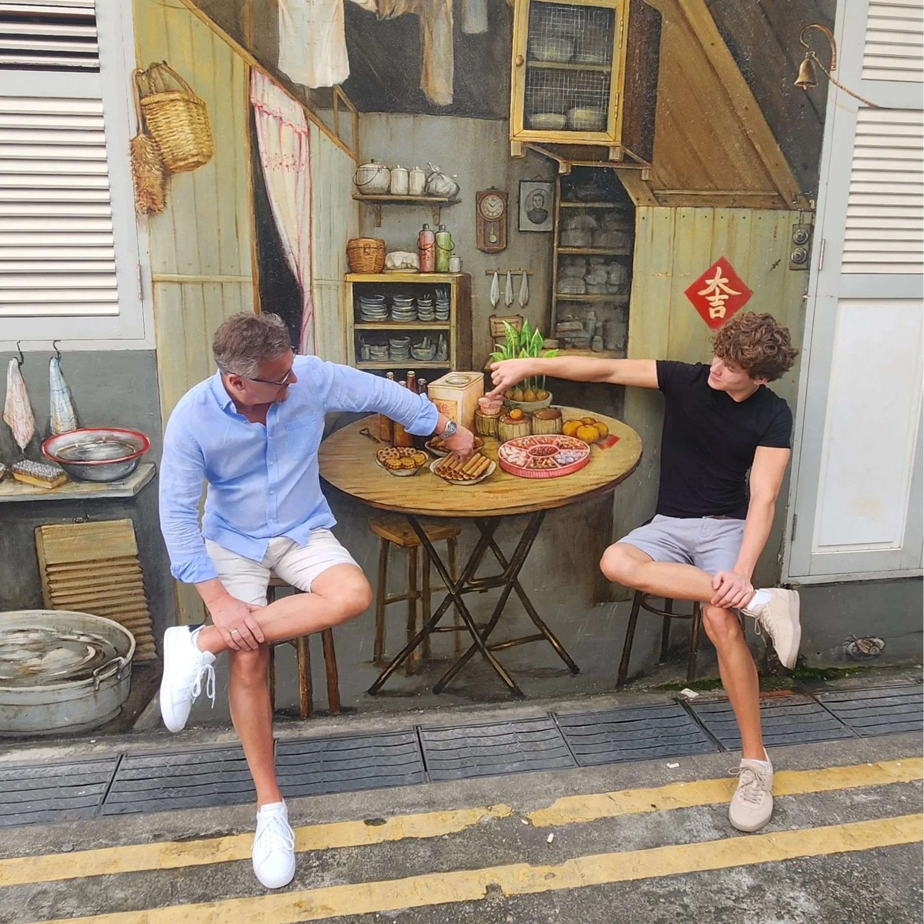 Two young men with curly hair and wearing casual clothes are sitting at a small round wooden table outside, engaging in a playful arm wrestling match. One man is in a black t-shirt and beige shorts, while the other is wearing a light blue shirt and w