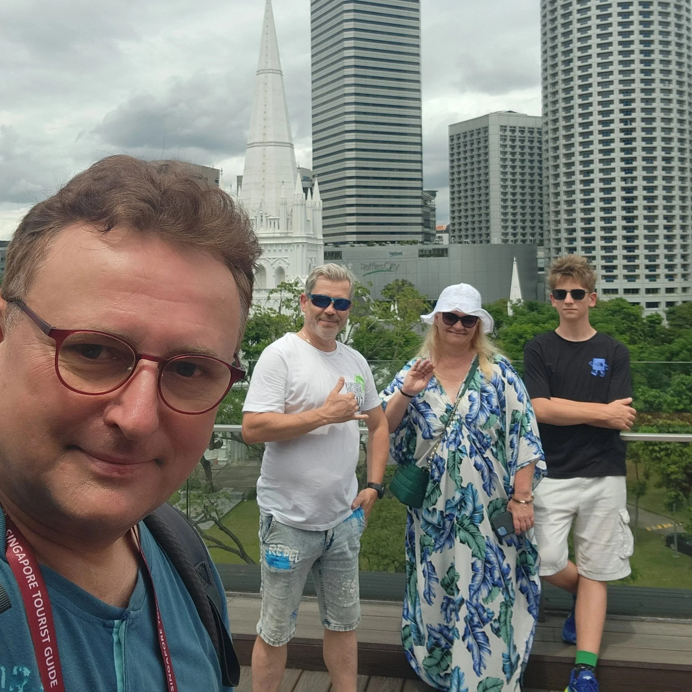 A group of people standing outdoors with high-rise buildings in the background, some wearing sunglasses and casual clothing, with the front man partially taking a selfie.
