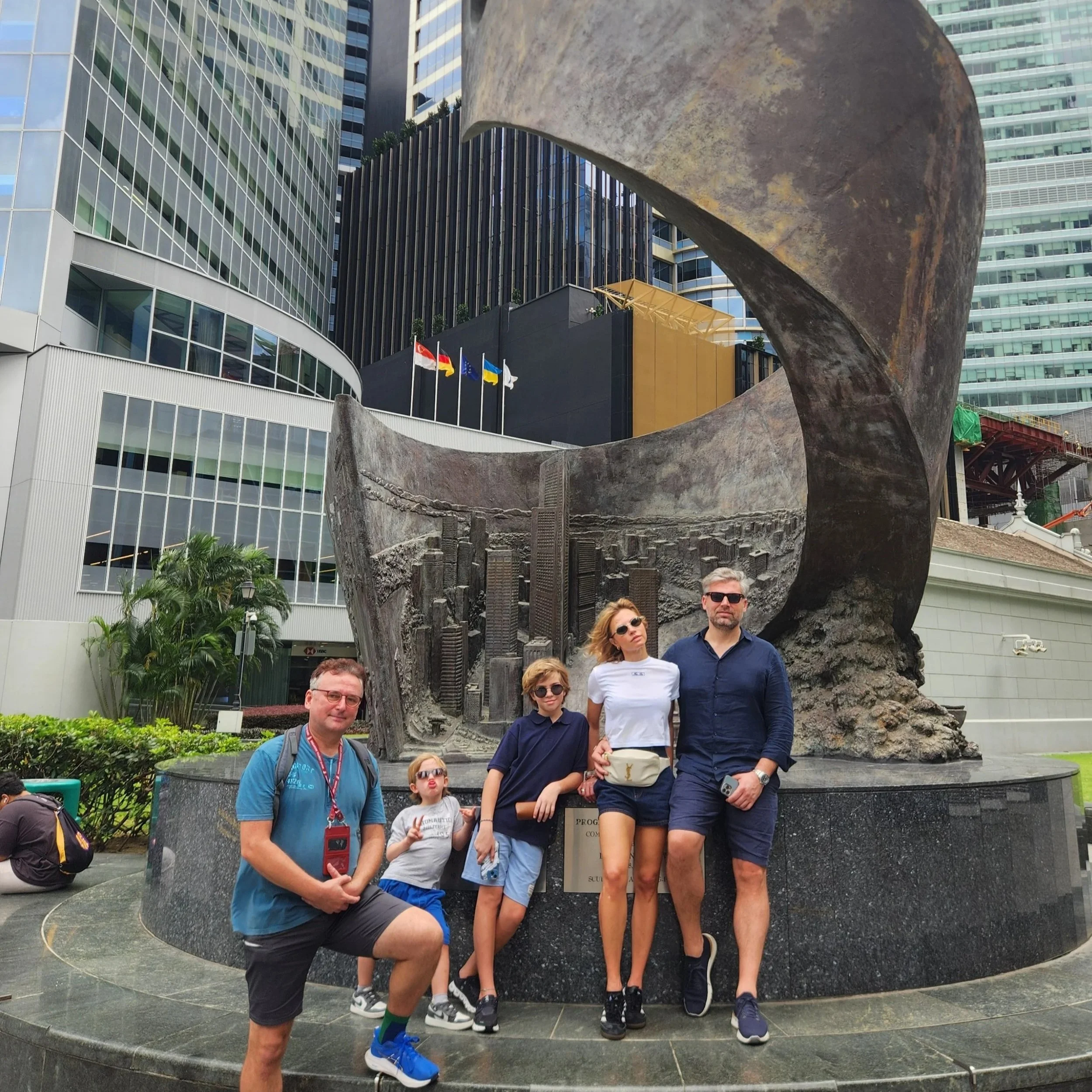 A group of six tourists posing in front of a large, abstract metal sculpture in an urban setting. City buildings surround the area, and four flags are visible behind the sculpture.