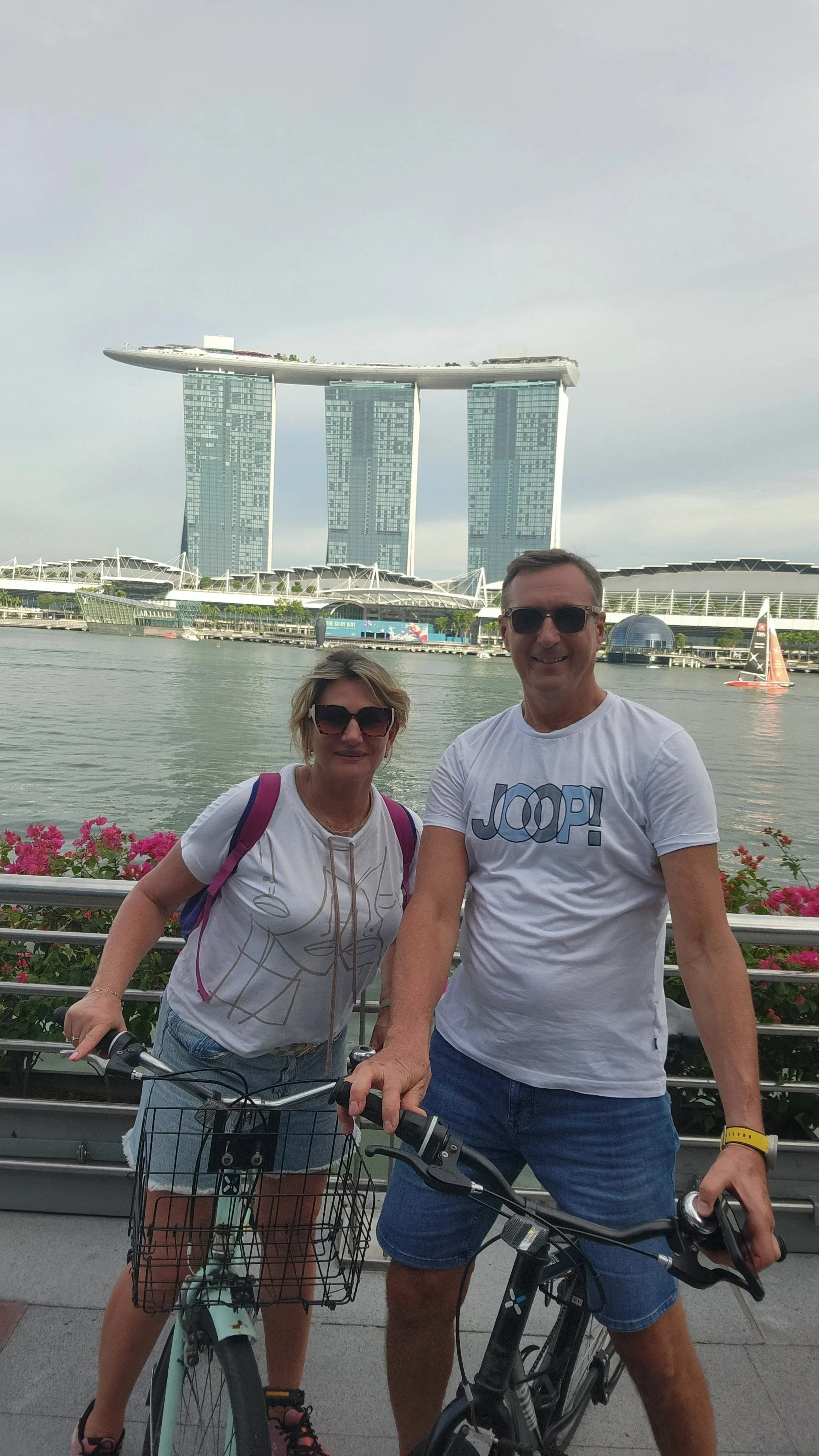 A man and a woman posing with bicycles in front of Marina Bay Sands hotel in Singapore, with a waterway and sail-shaped structure in the background.