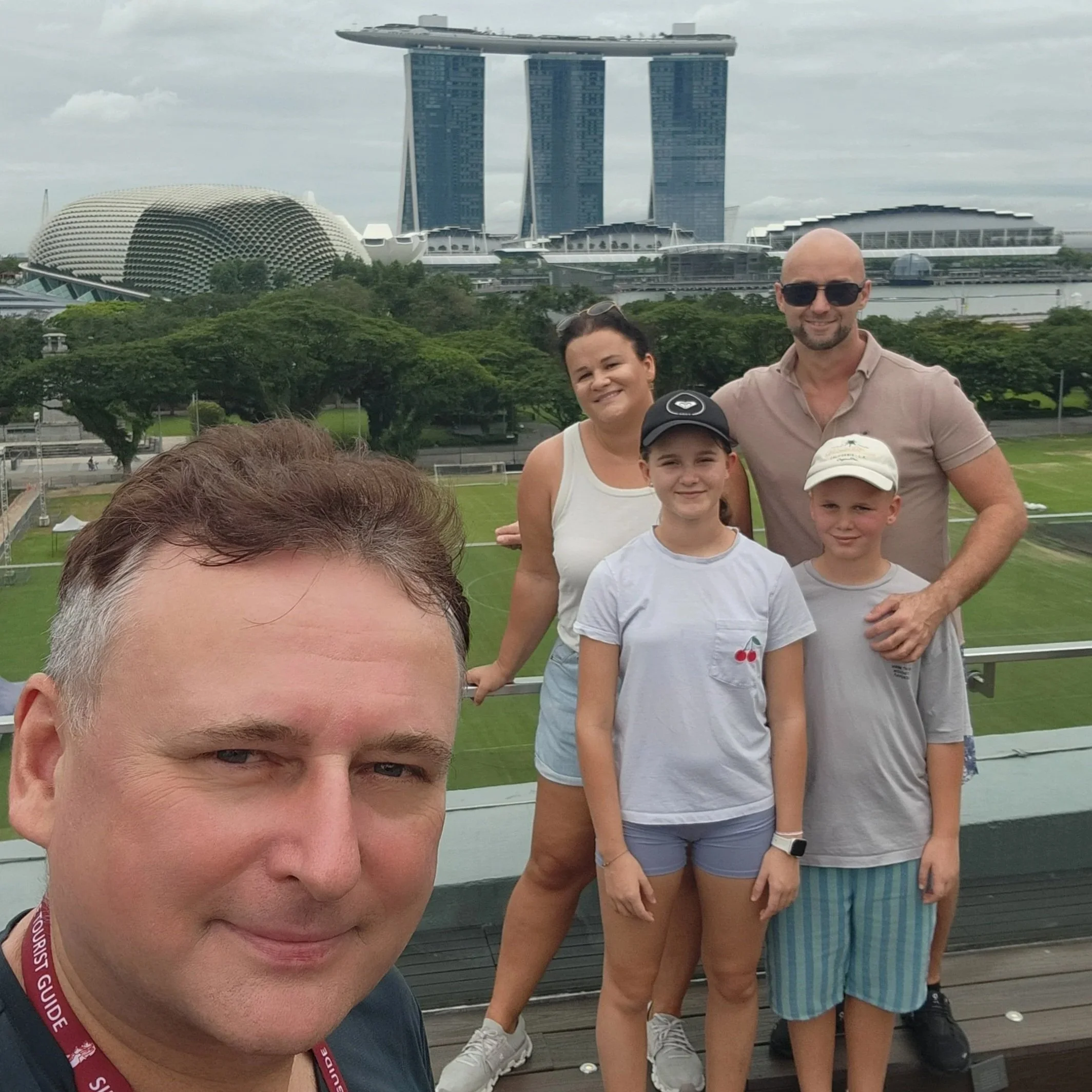 A group of people, including a man with sunglasses and two children, standing on a wooden deck with greenery around, with the Marina Bay Sands hotel and the ArtScience Museum in Singapore visible in the background.