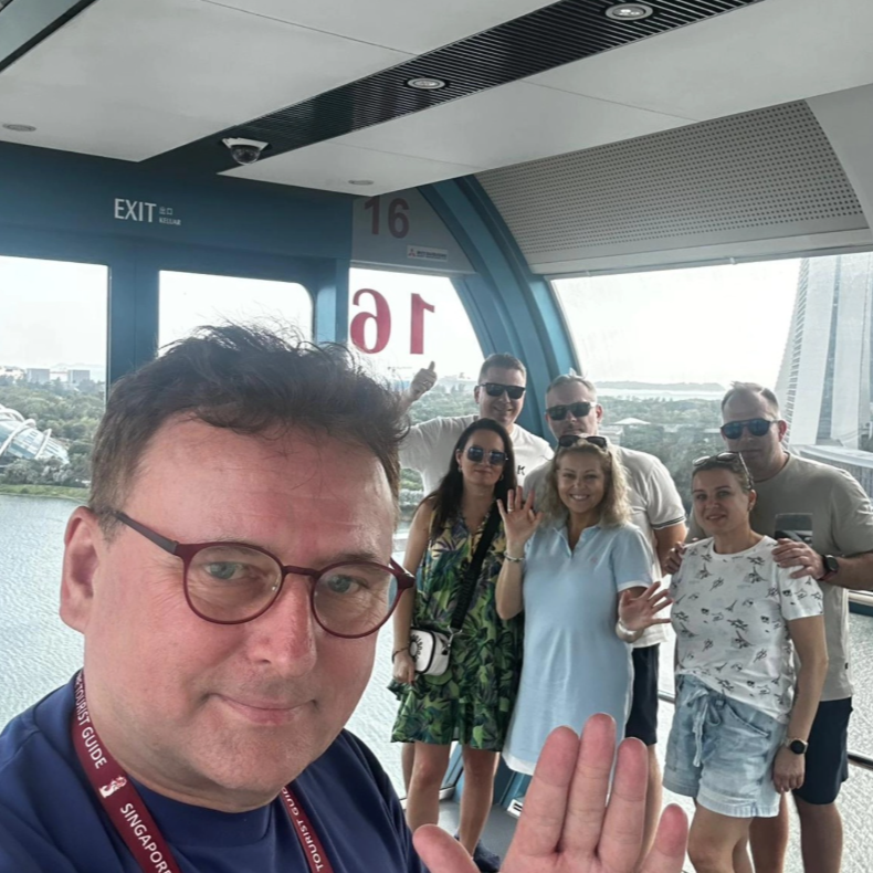 Group of seven people posing for a photo inside a Ferris wheel cabin with a city and river view in the background. One person is taking a selfie while the others wave.