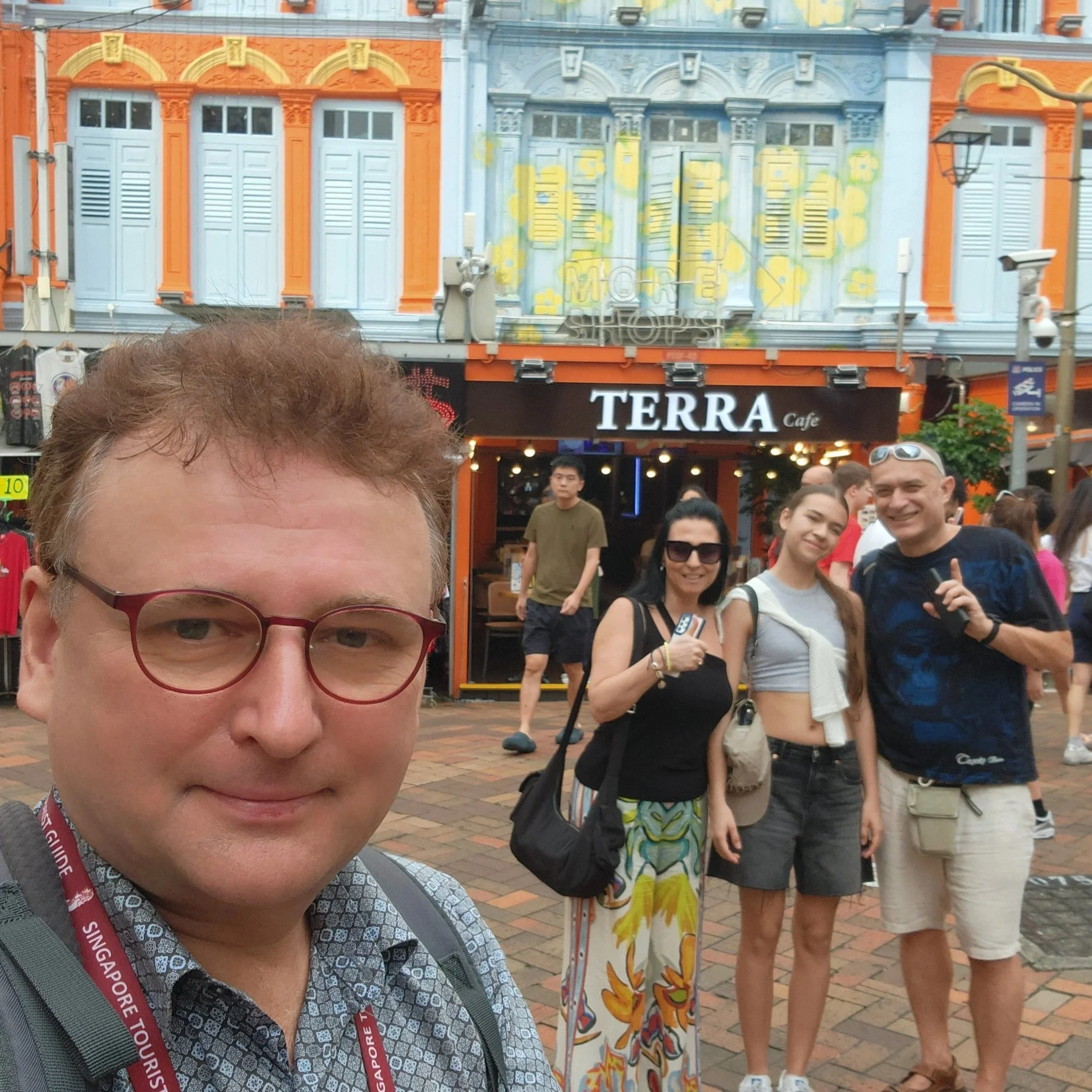 A man with glasses and a backpack taking a selfie on a busy street with people and colorful storefronts in the background.