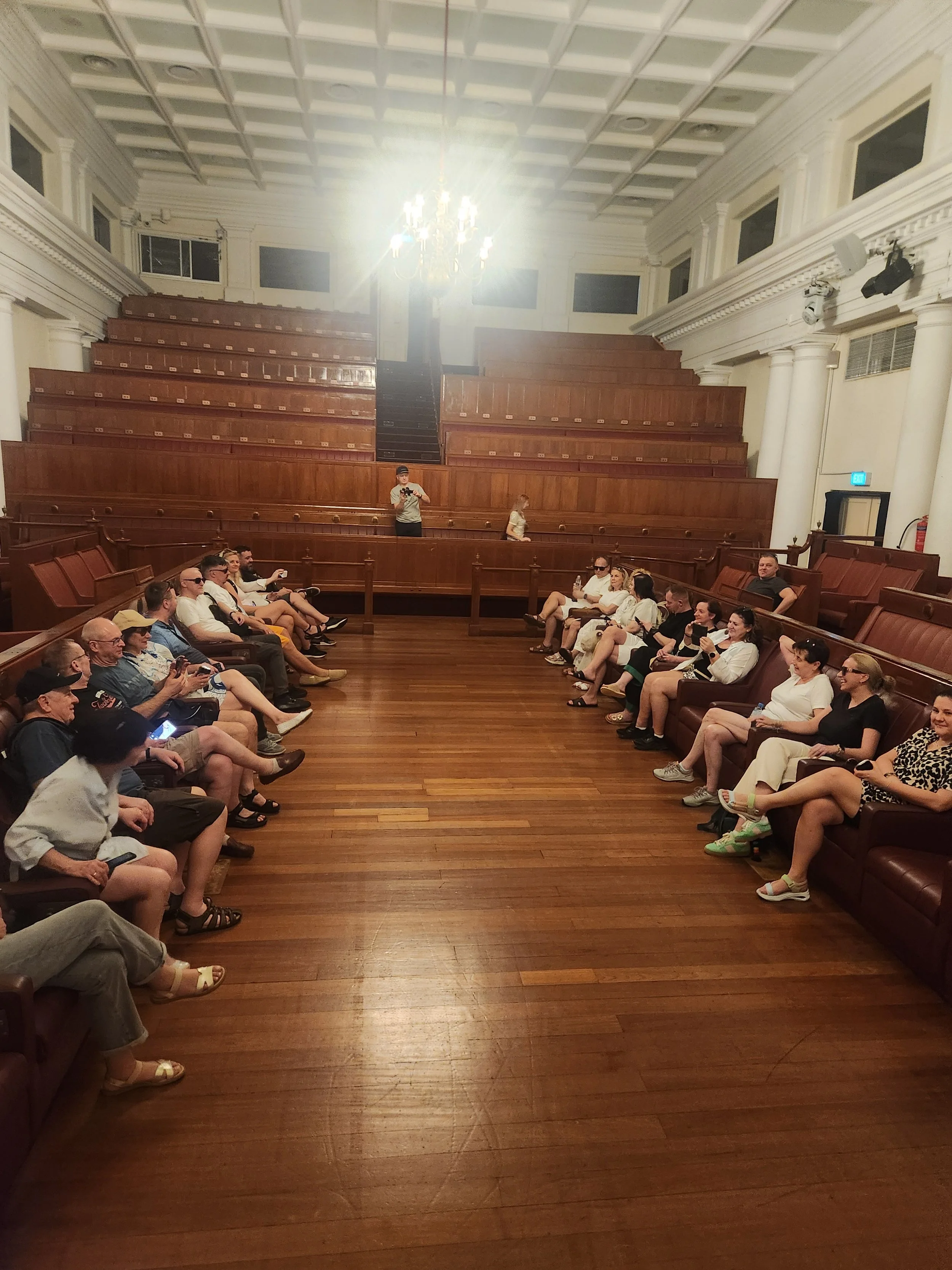 A group of people sitting in a semi-circle in a wood-paneled room with a high ceiling, chandeliers, and large pillars. Some are wearing sunglasses and casual clothes, and they are engaged in conversation or using their phones.