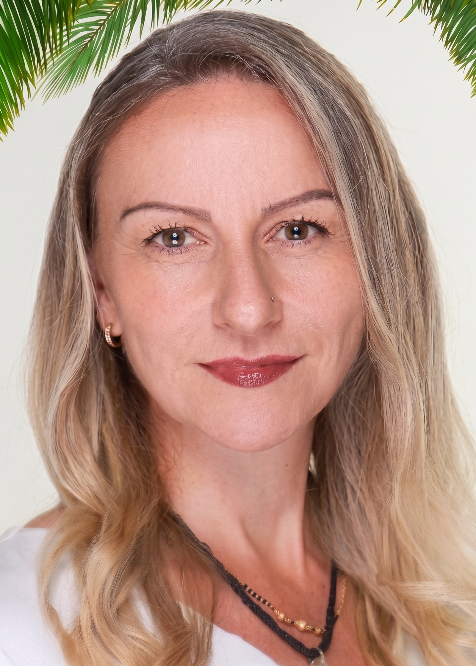 A close-up photo of a woman with light skin, long wavy blonde hair, and brown eyes, smiling slightly against a plain light background, with some green palm leaves overlaying the top corners of the image.