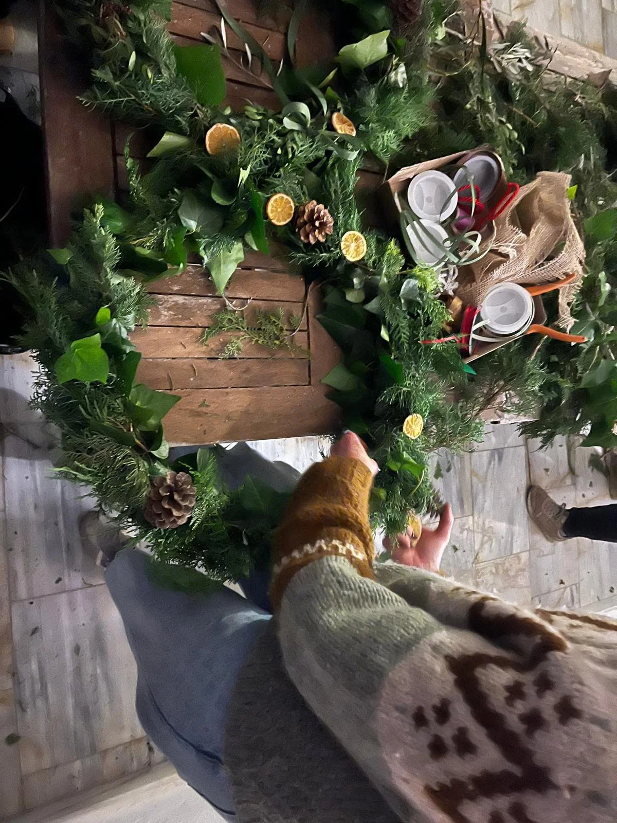 Person arranging a festive holiday wreath with greenery, pine cones, and dried orange slices on a wooden table, surrounded by holiday decorations and supplies.