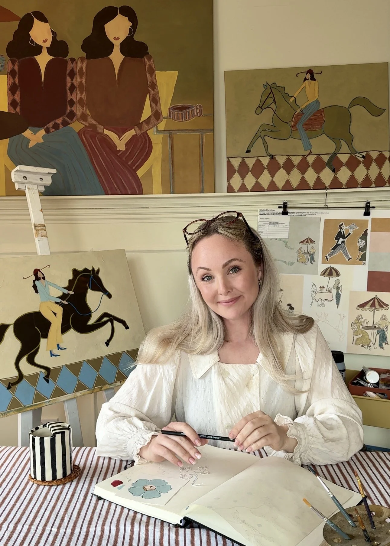 A woman with blonde hair and glasses on her head, wearing a cream-colored blouse, sits at a table with art supplies. She is holding a pen and appears to be sketching. Behind her are paintings of women and men riding horses, displayed on the wall and leaning on easels, with art tools and sketches visible on the table and wall.