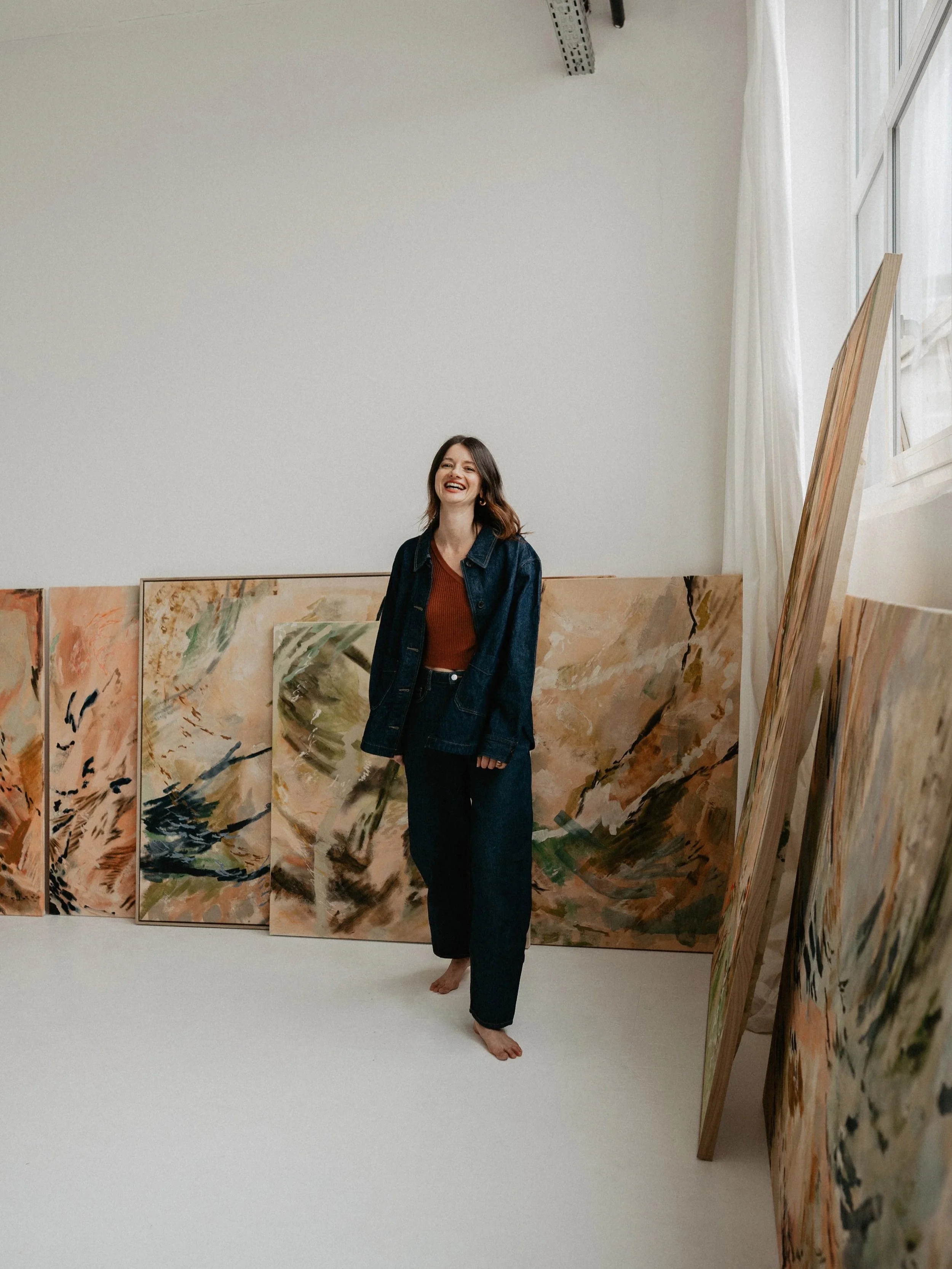 A woman with long brown hair, wearing a denim jacket and dark pants, standing barefoot in an art gallery with colorful abstract paintings behind her.