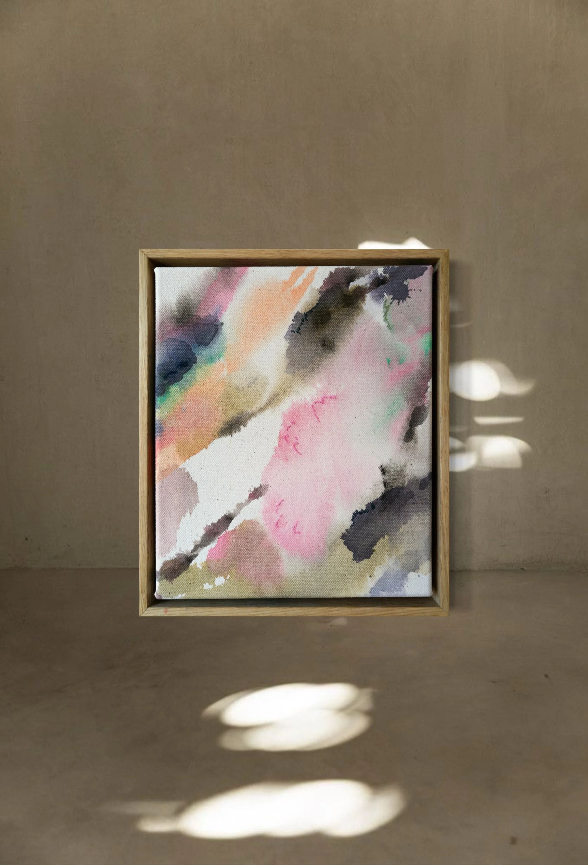 Small abstract watercolor painting in a wooden frame, featuring pink, black, green, and blue colors, on a beige surface with sunlight and shadow.