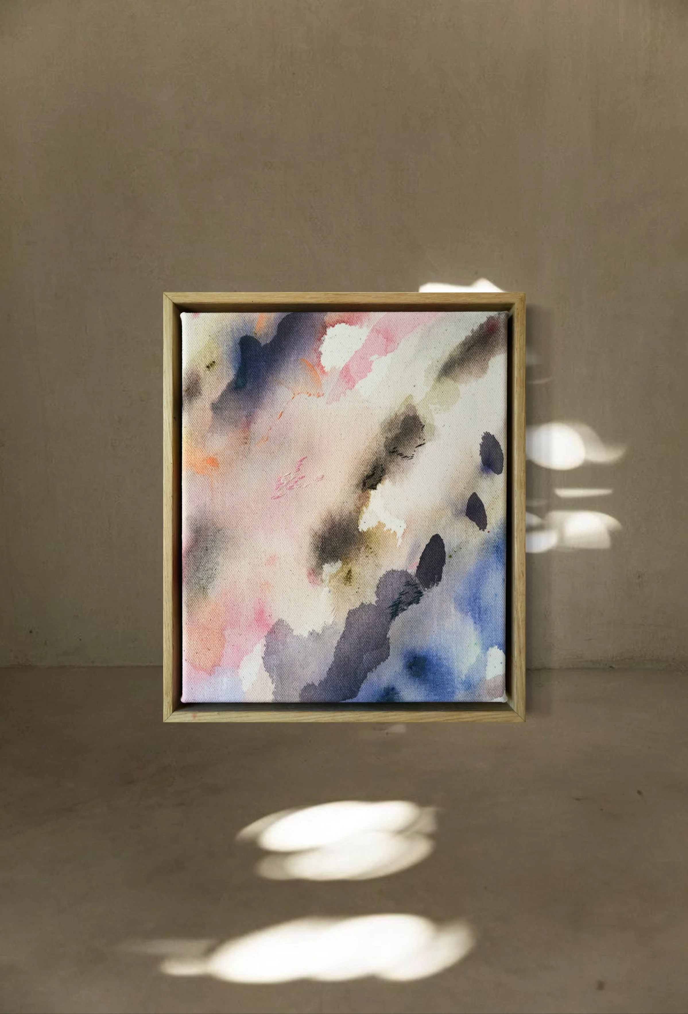 A framed abstract watercolor painting with soft pastel tones on a beige wall, illuminated by natural light creating shadow patterns on the wall.