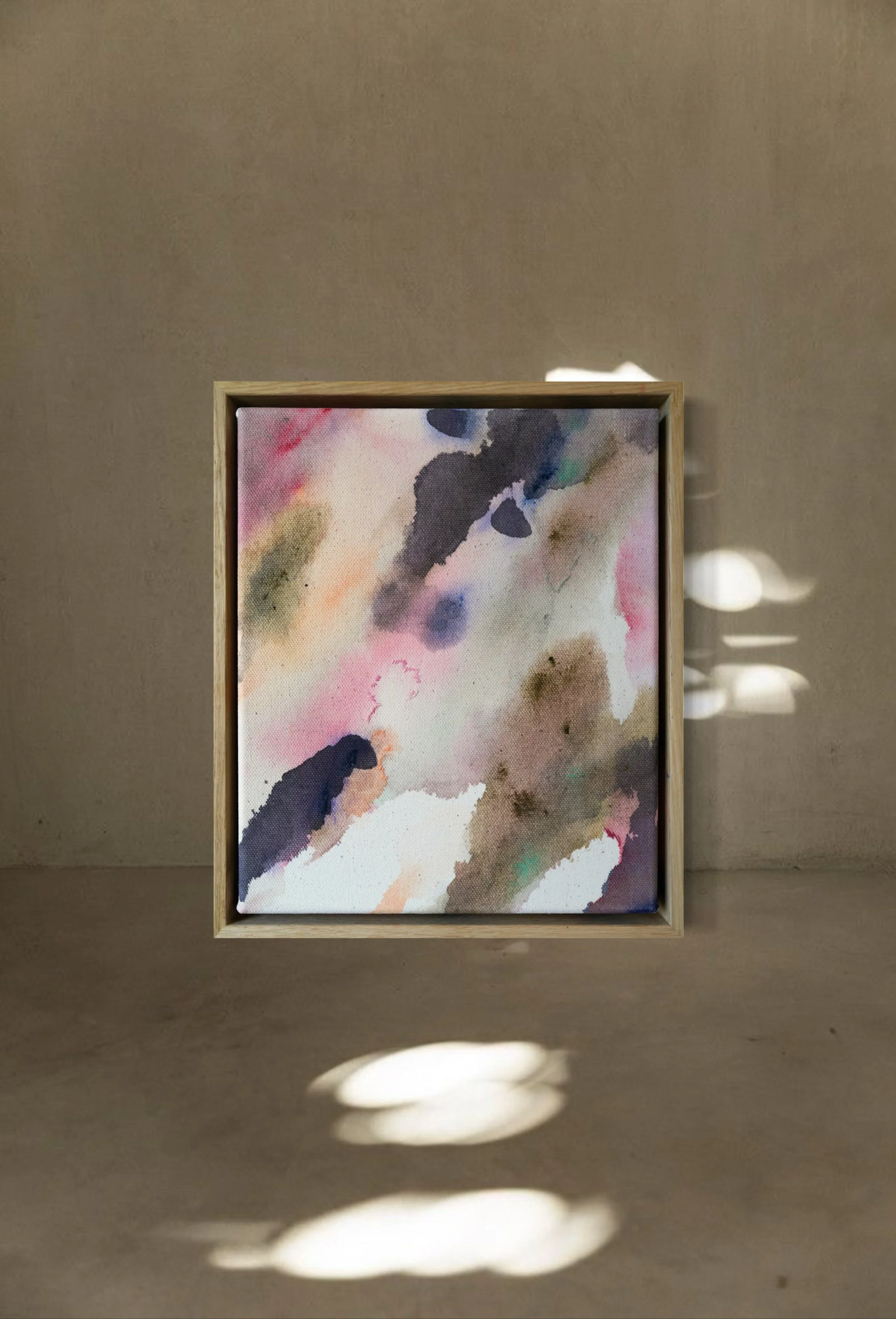 Abstract watercolor painting in a light wooden frame, displayed on a beige wall with light and shadow patterns.
