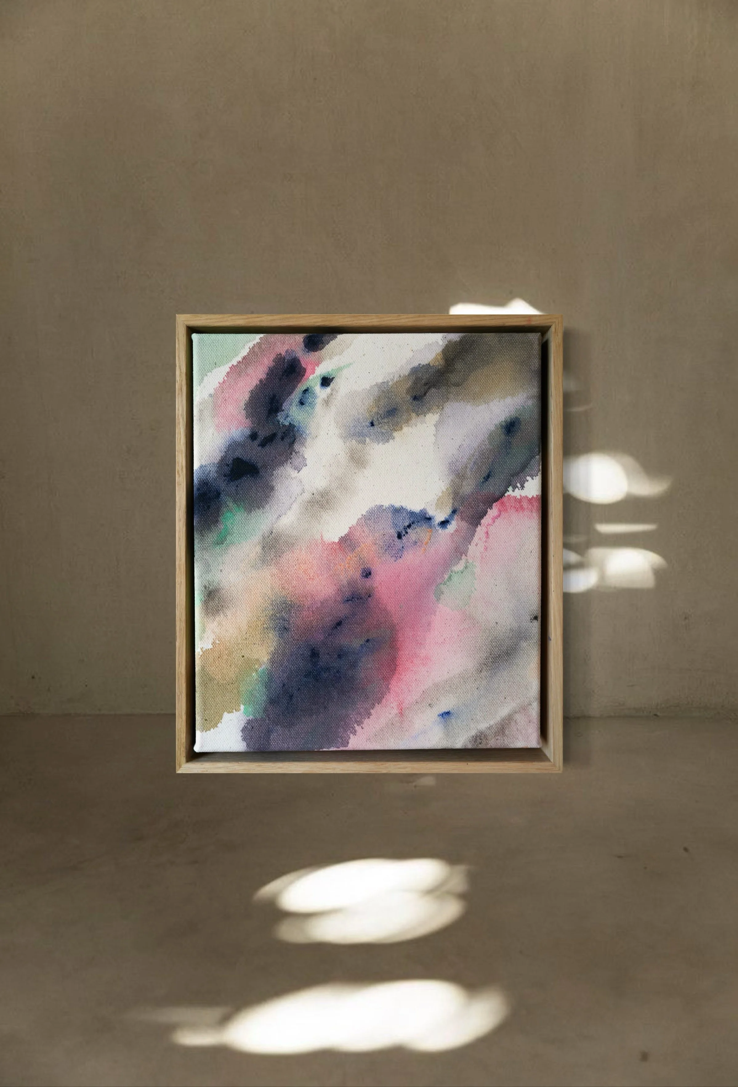 Abstract watercolor painting with pink, purple, green, and blue hues in a wooden frame, hung on a beige wall, with light reflections and shadows on the wall.