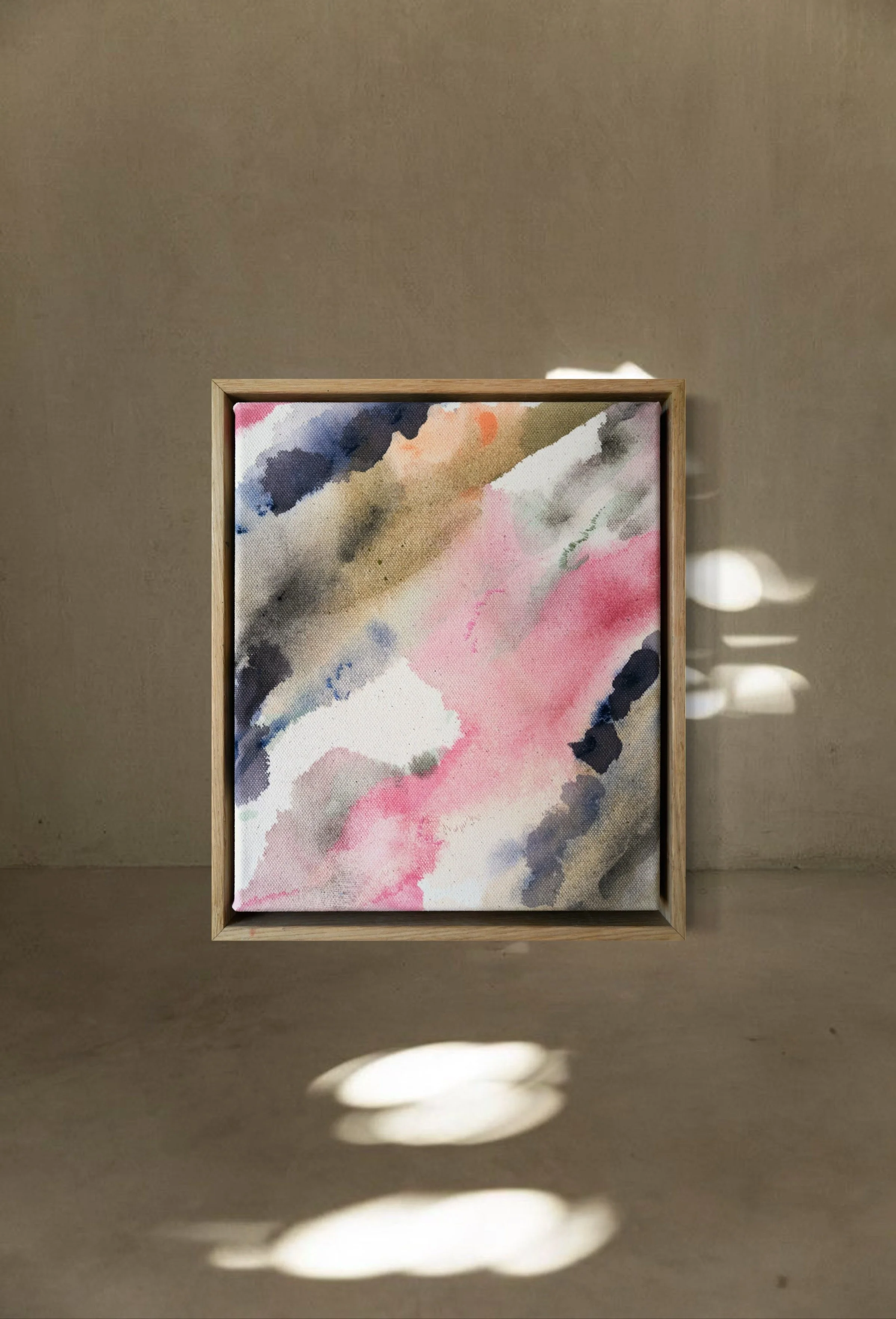 An abstract watercolor painting with pink, gray, black, beige, white, and blue colors in a wooden frame, placed on a concrete surface with shadows and reflections.