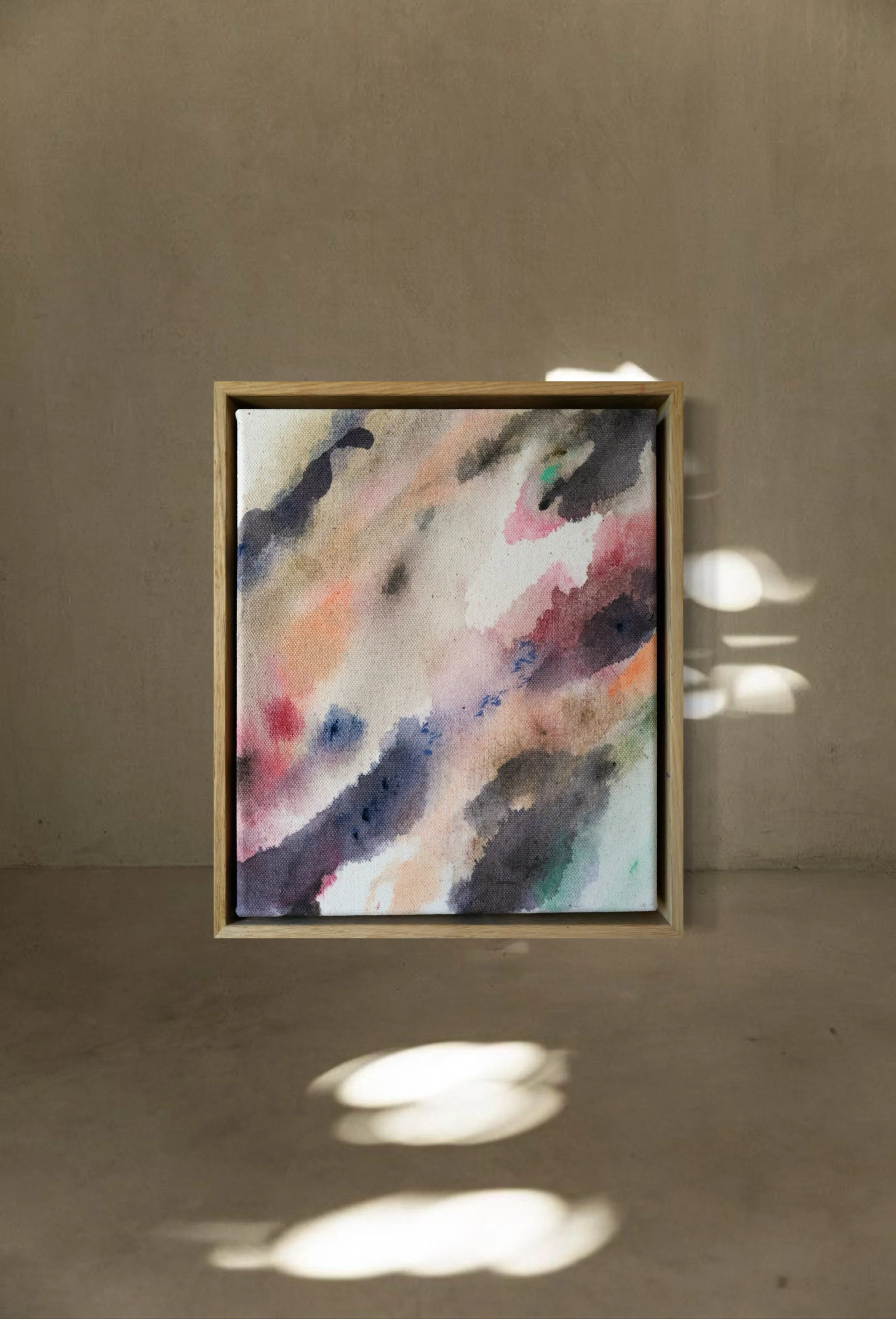 A framed abstract watercolor painting with soft pastel and dark colors, displayed on a beige wall. Light shadows are cast on the wall and floor.
