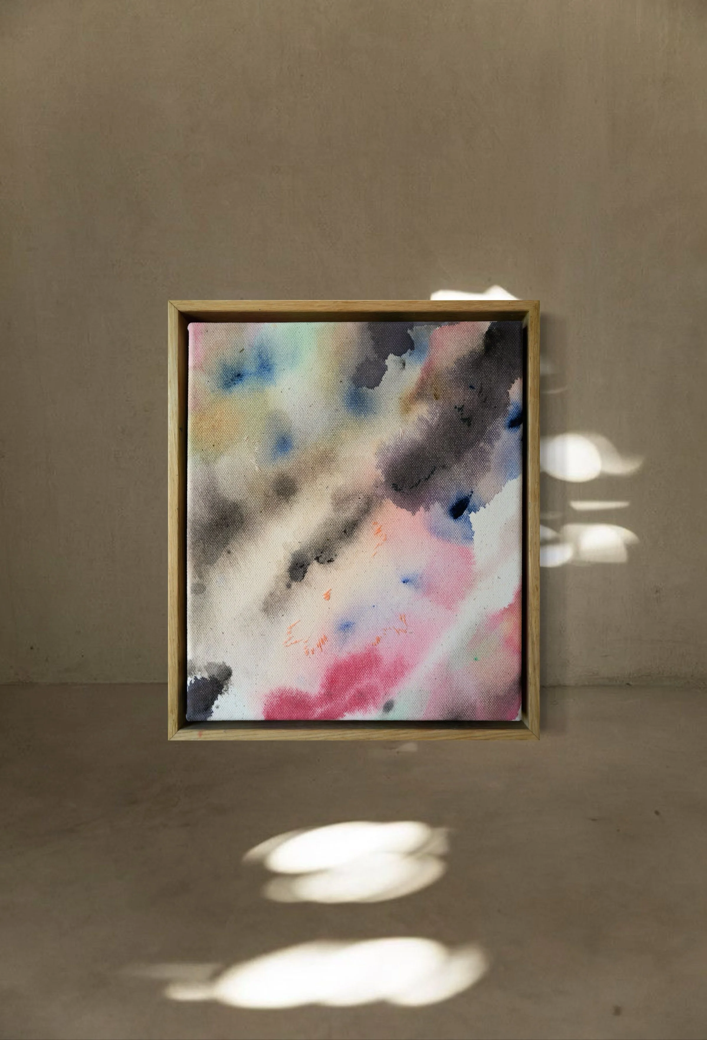 Abstract watercolor painting in a wooden frame with black, pink, blue, green, and yellow hues, set against a beige wall with shadows cast on the floor.