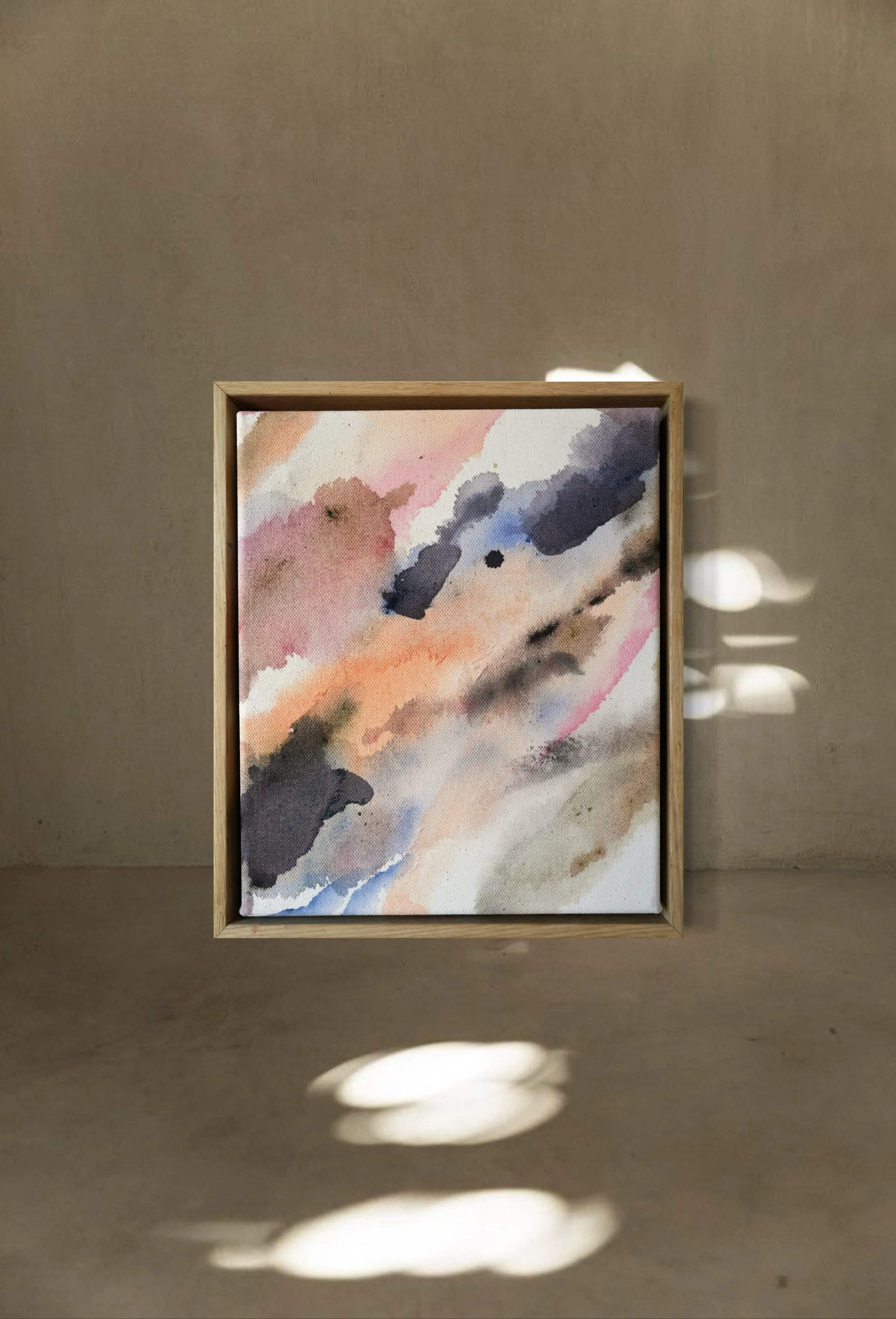 Abstract watercolor painting with soft pink, blue, black, and beige brush strokes in a wooden frame, displayed against a beige wall with light and shadow on the floor.
