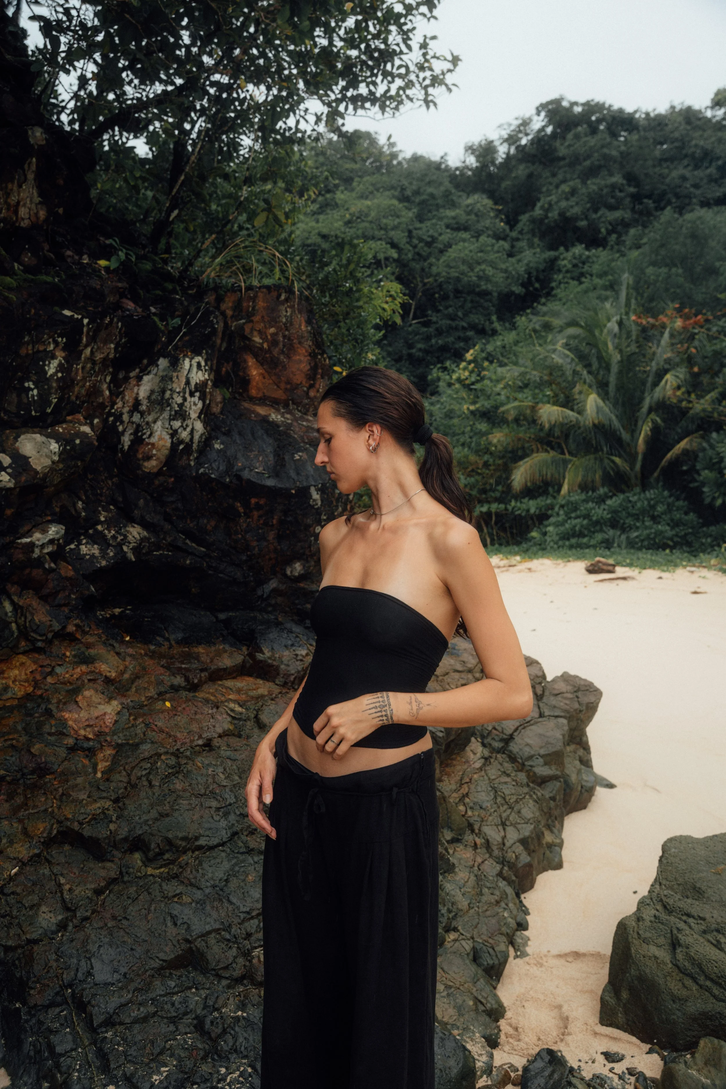 A woman with dark hair tied back, wearing a strapless black top and black pants, standing outdoors near rocks and sandy beach, with lush green trees in the background.