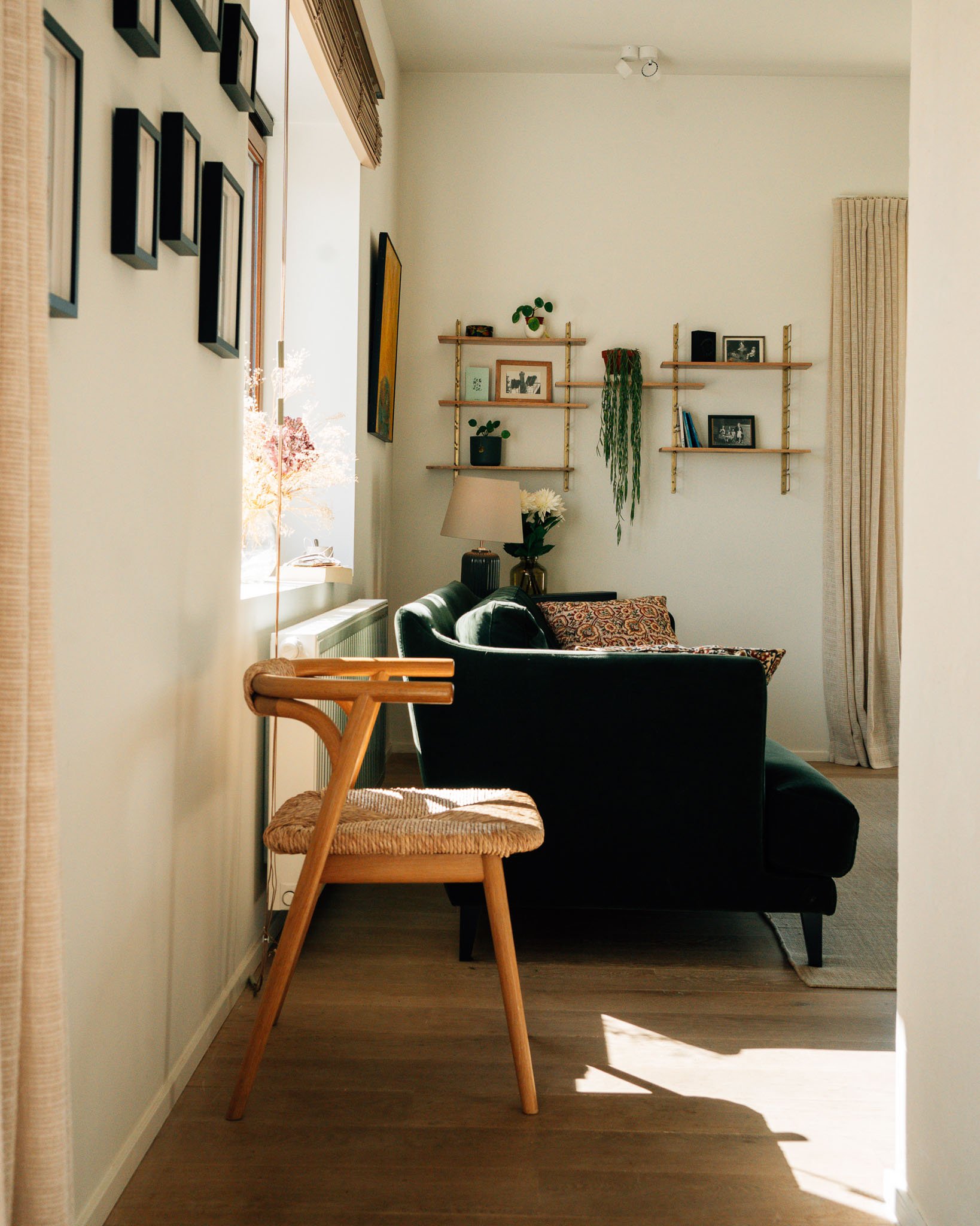 A cozy living room corner with a black velvet sofa, patterned cushion, beige curtains, a wooden coffee table, a wall with floating shelves displaying framed photos and potted plants, a table lamp, and sunlight creating shadows on the wooden floor.