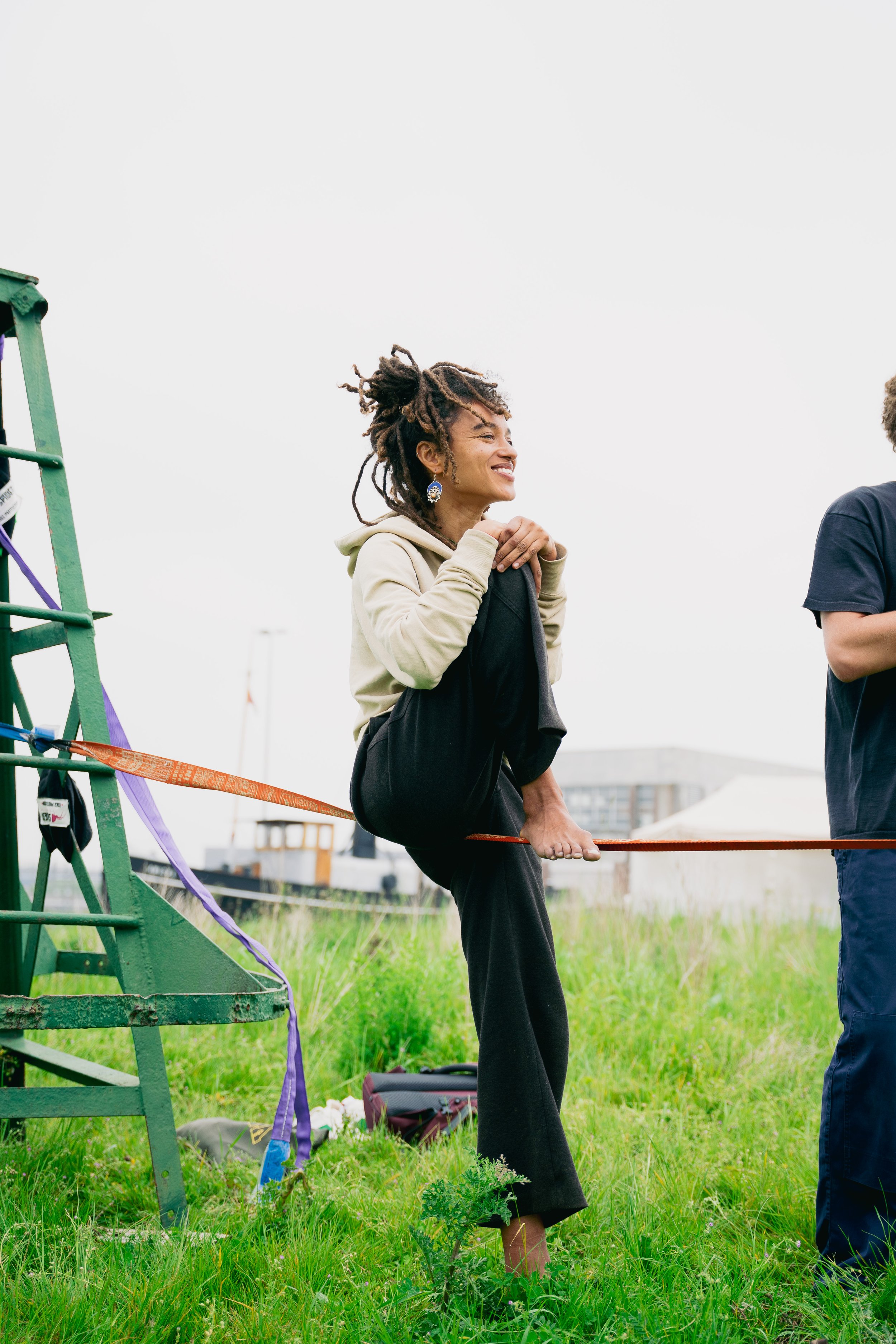 A woman with dreadlocks smiling and sitting on a slackline anchored to a green structure, outdoors on grassy ground in a cloudy environment.