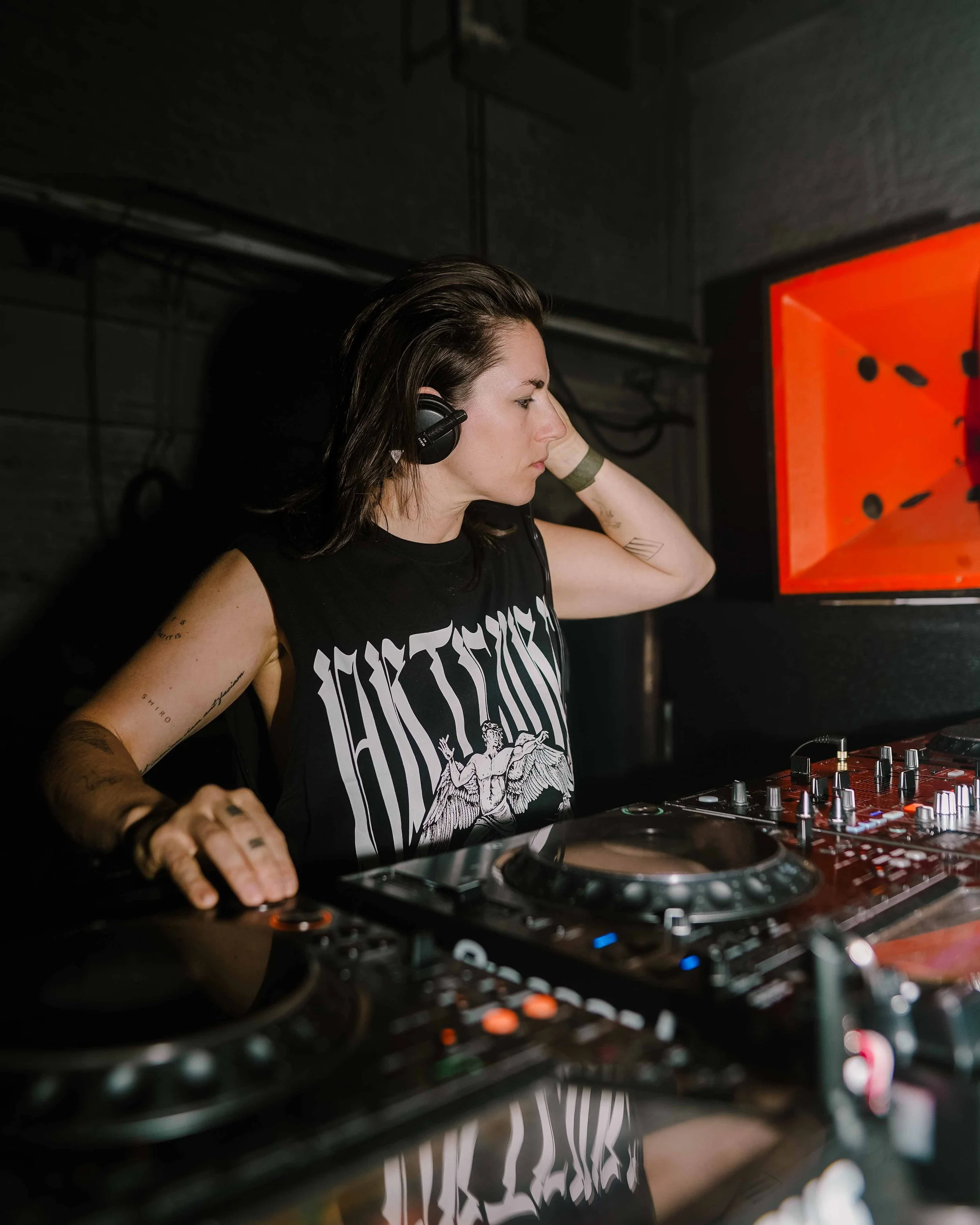 A female DJ with long dark hair and tattoos on her arms, wearing headphones, adjusting music equipment in a dimly lit setting with a red and orange background.