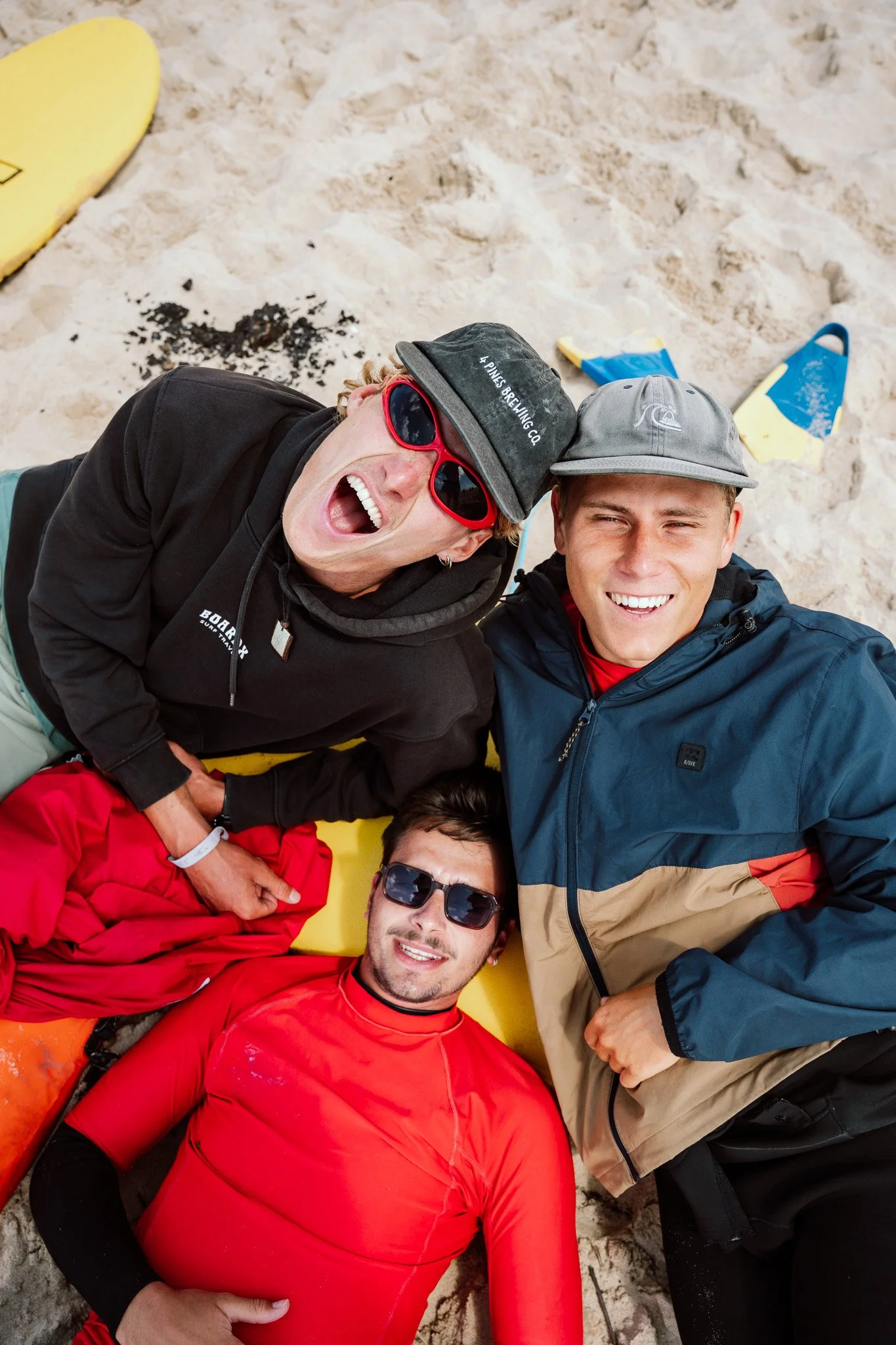 Four people lying on the sand at the beach, smiling and looking up at the camera. Two men are wearing sunglasses, one woman is wearing red sunglasses and a black hoodie, and another person in a red jacket is lying beneath them. There are some blue an