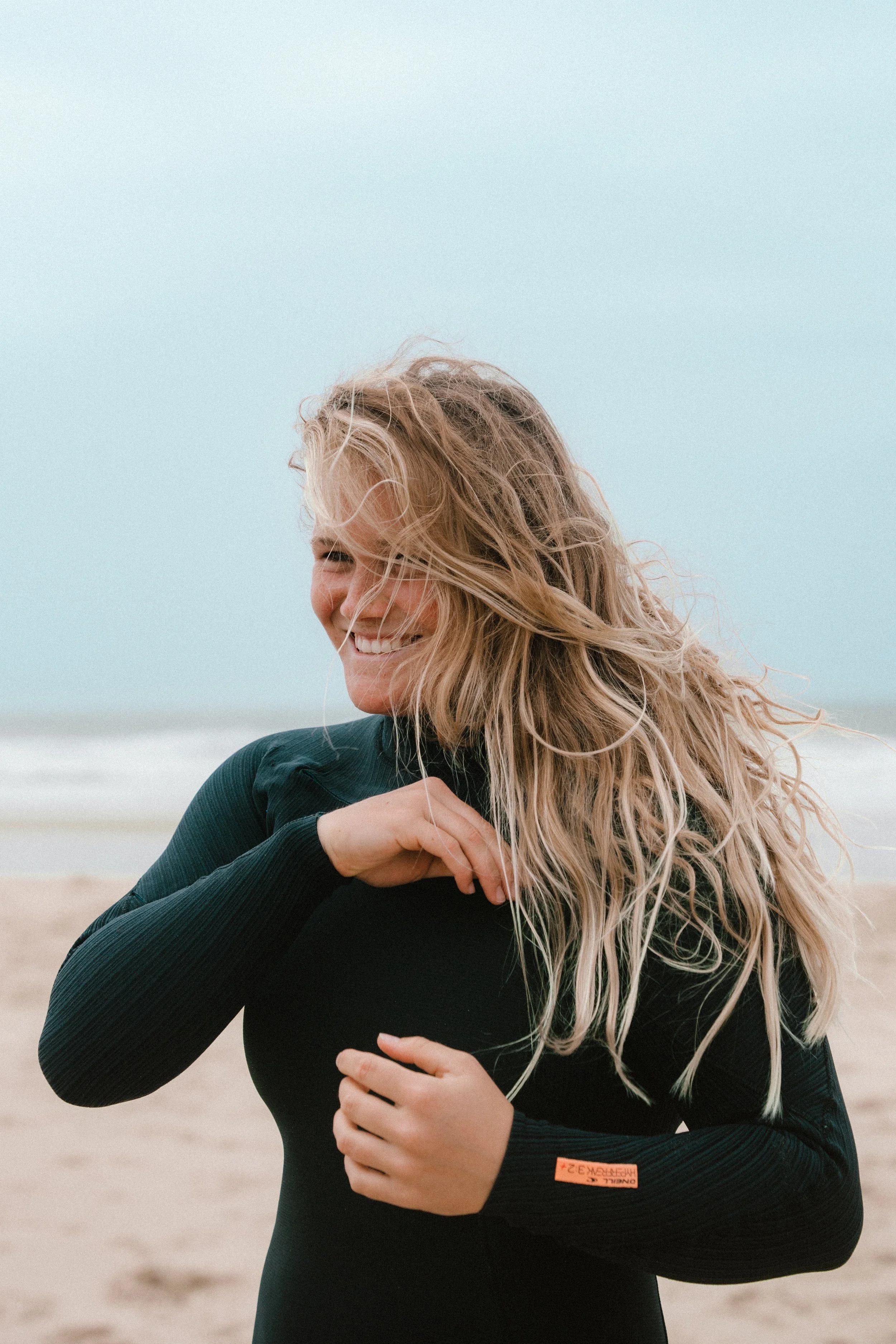 A woman smiling and laughing at the beach, with sandy dunes and a light blue sky in the background, wearing a black long-sleeve shirt.