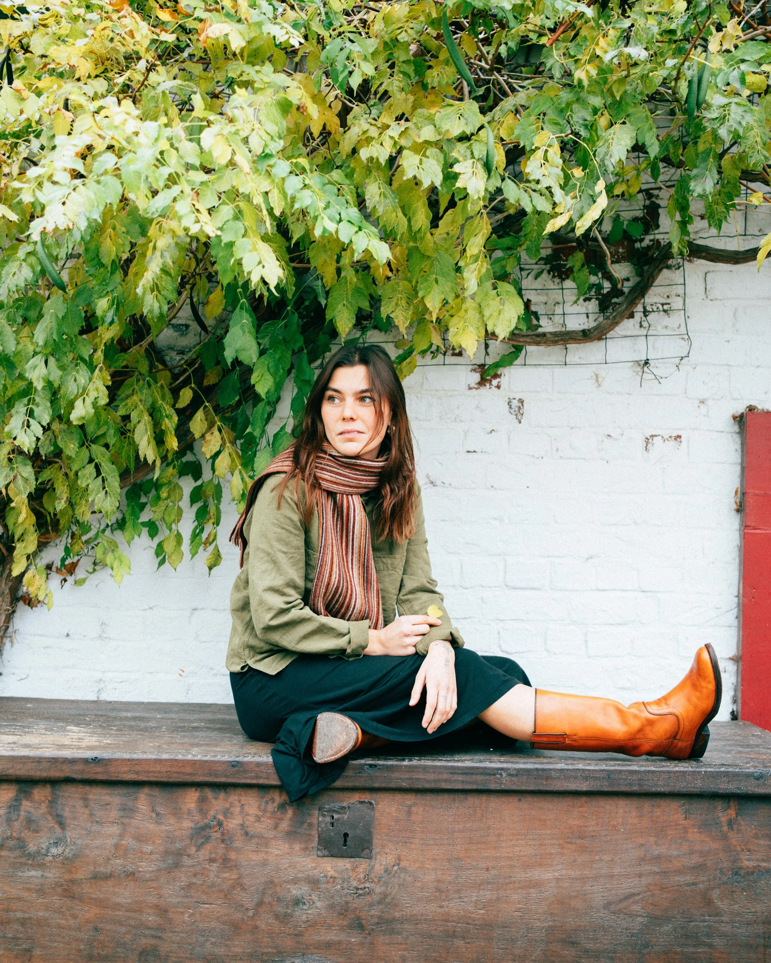 A woman with brown hair and fair skin sitting on a wooden bench outdoors against a white brick wall with green leafy vine plants.