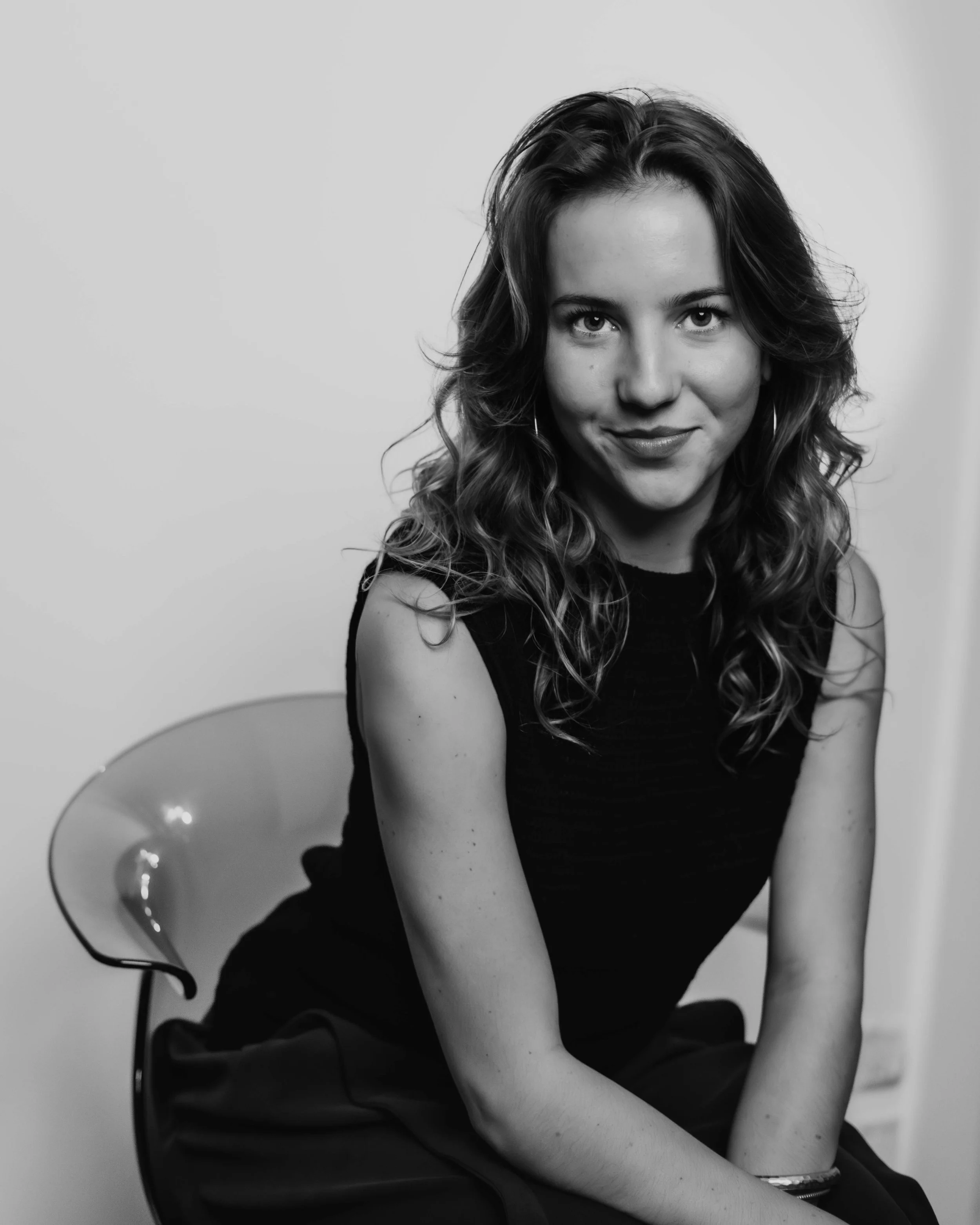 Black and white photo of a young woman with wavy hair, sitting on a chair, looking directly at the camera with a slight smile. studio setting