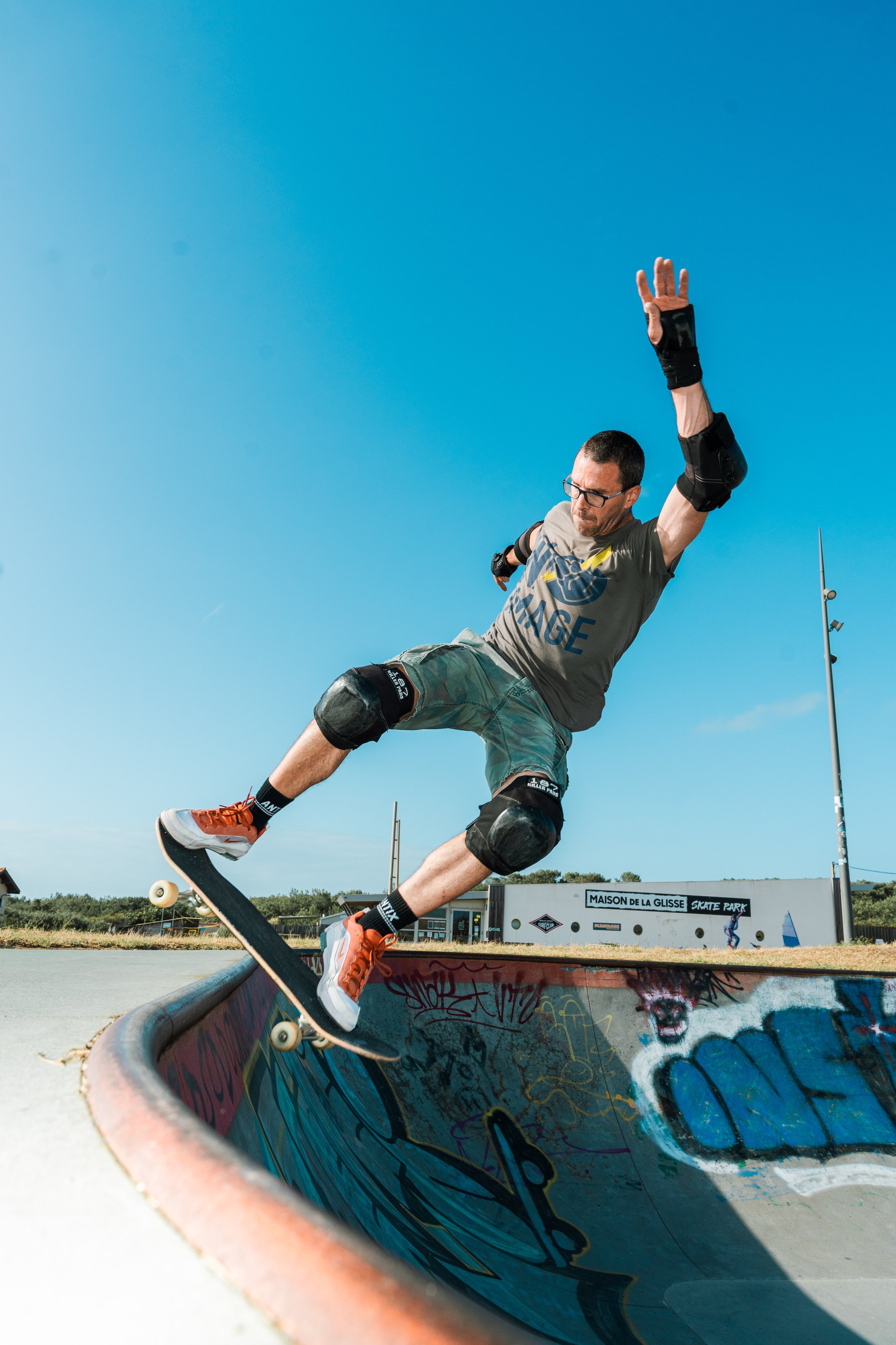 Skateboarder in a skate park performing a trick on a bar, with graffiti on the skate bowl, wearing protective gear, under a clear blue sky.