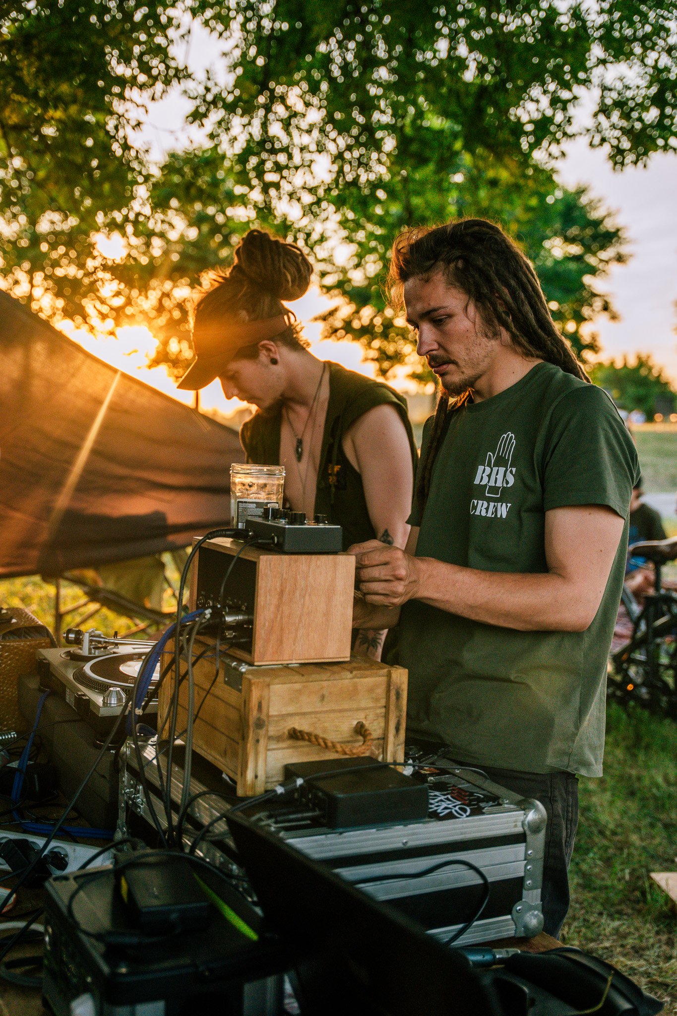 Two young men with dreadlocks working with electronic music equipment outdoors at sunset, with trees and a sunset in the background.