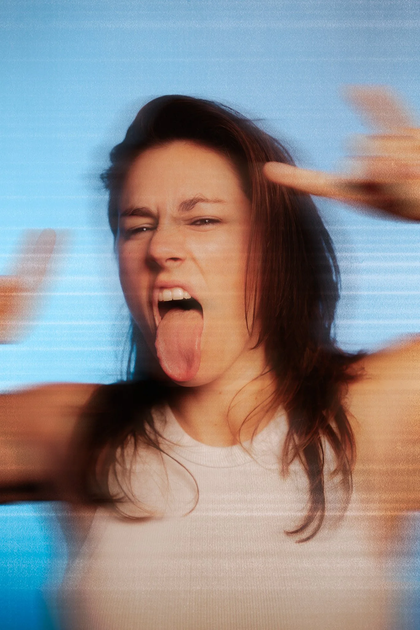 A woman with dark hair and light skin sticking out her tongue and making a peace sign with her fingers, with an expression of excitement or playfulness.
