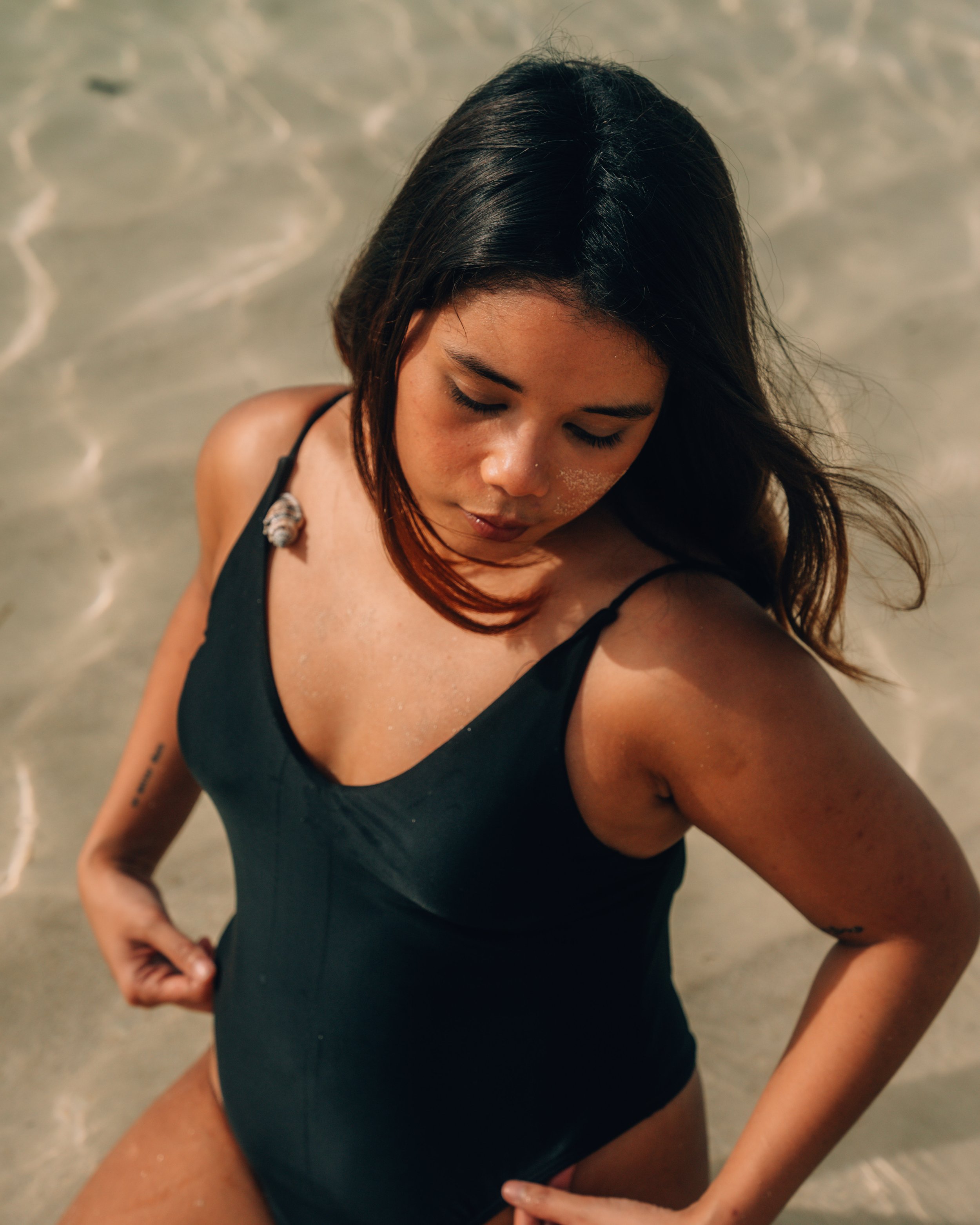 A woman with dark hair in a black swimsuit kneeling on sandy beach with water in background, eyes closed, head slightly tilted downward.