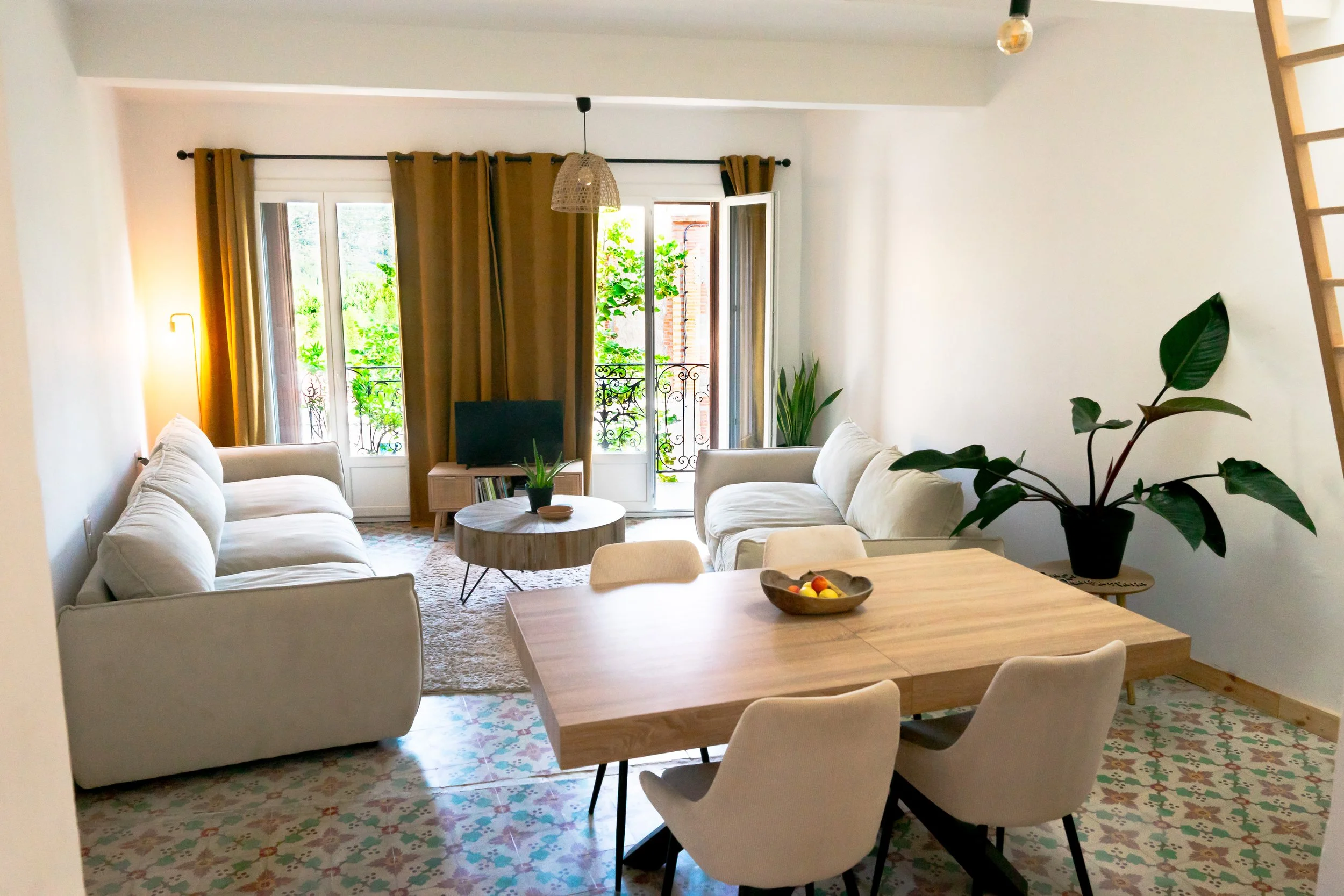 Living room with white couches, a round wooden coffee table, large open windows with brown curtains, potted plants, a small TV, and a dining table with chairs. Bright sunlight streams in.