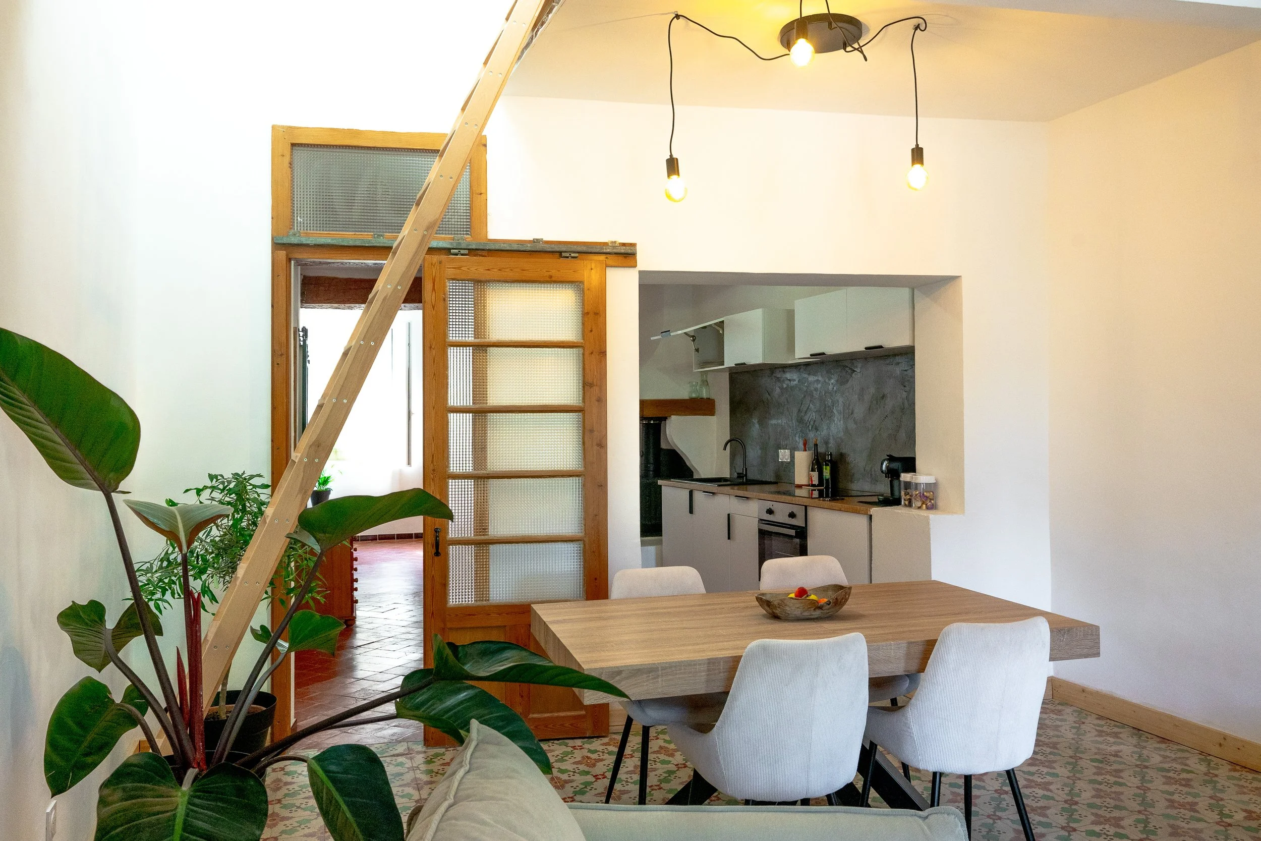 Interior view of a modern kitchen and dining area with white walls, a wooden dining table, white chairs, green plants, and hanging light bulbs.