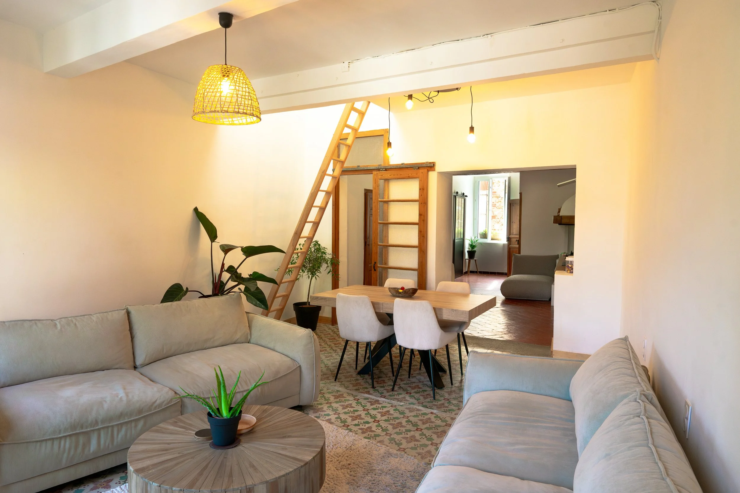 Living room featuring white sofas, a round wooden coffee table with a potted plant, a dining table with white chairs, a patterned rug, and an open background with a staircase leading to an upper level, natural light from a window, and warm hanging li