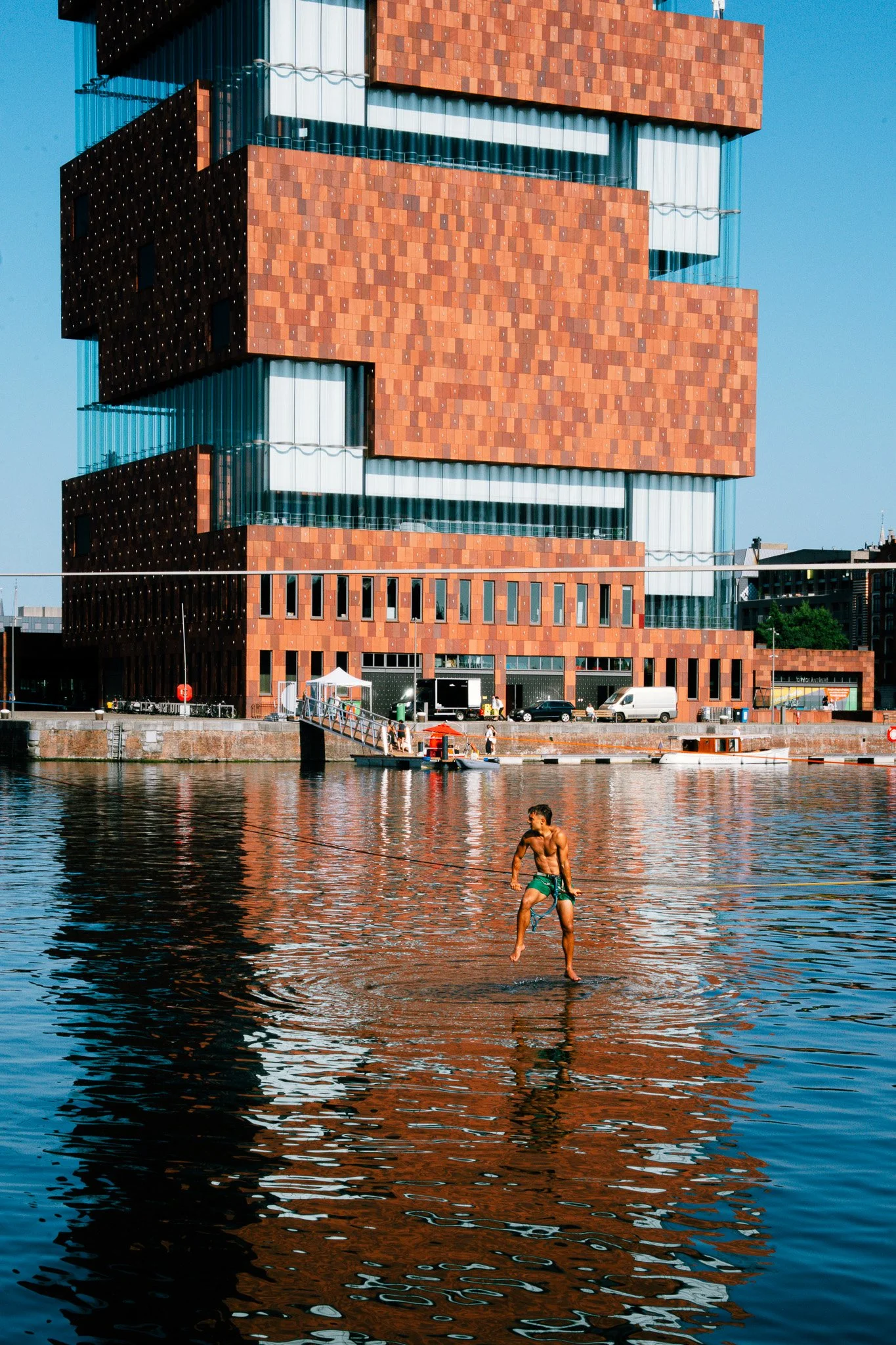 A man in green swim trunks stands in water, holding a leash attached to a dog, near a city waterfront with a modern, multi-story, red brick and glass building in the background.