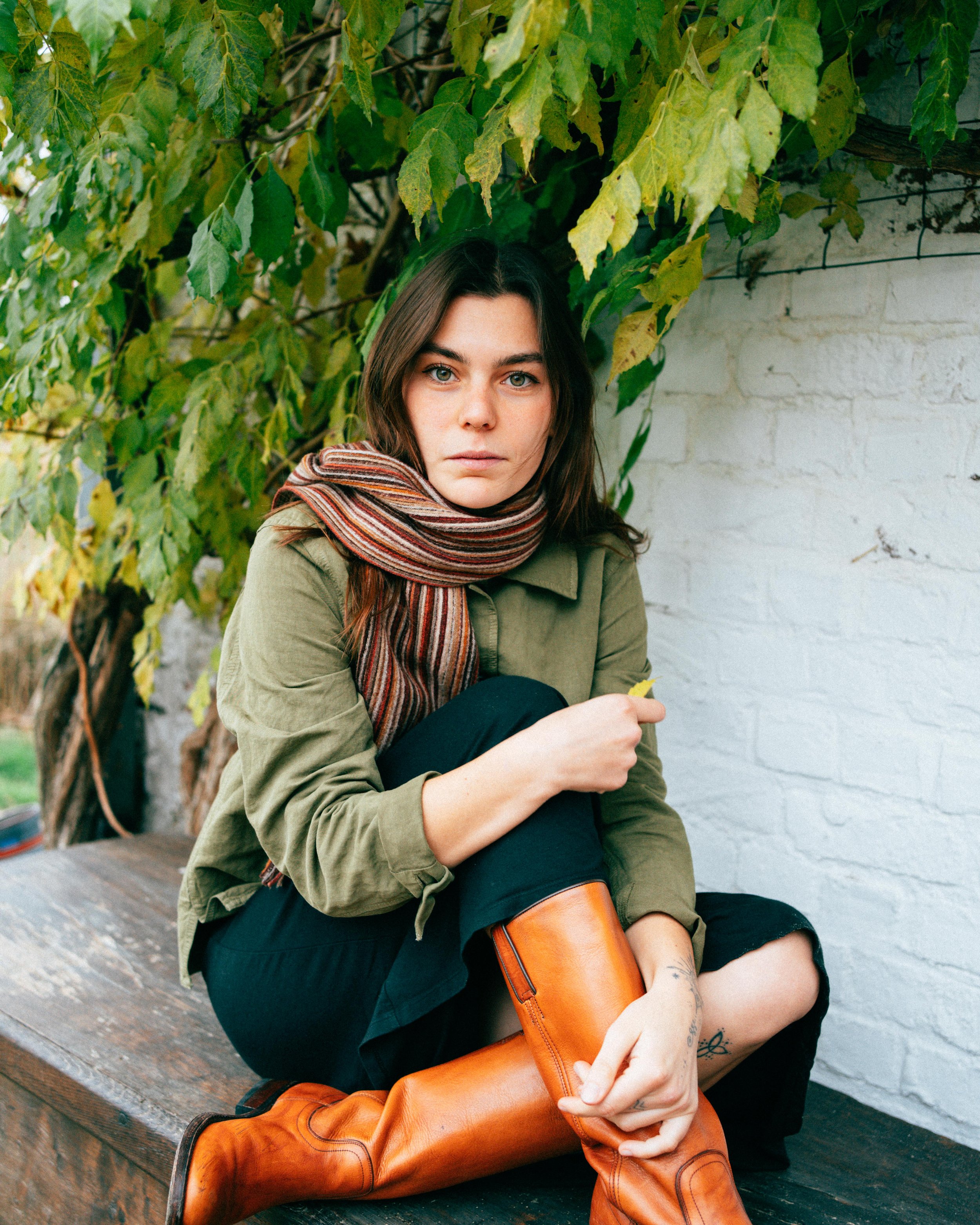 A young woman with long brown hair and light-colored eyes sitting on a wooden surface outside, surrounded by green leafy vines, wearing a tan jacket, striped scarf, black pants, and brown leather boots. She has tattoos on her left ankle and right wri