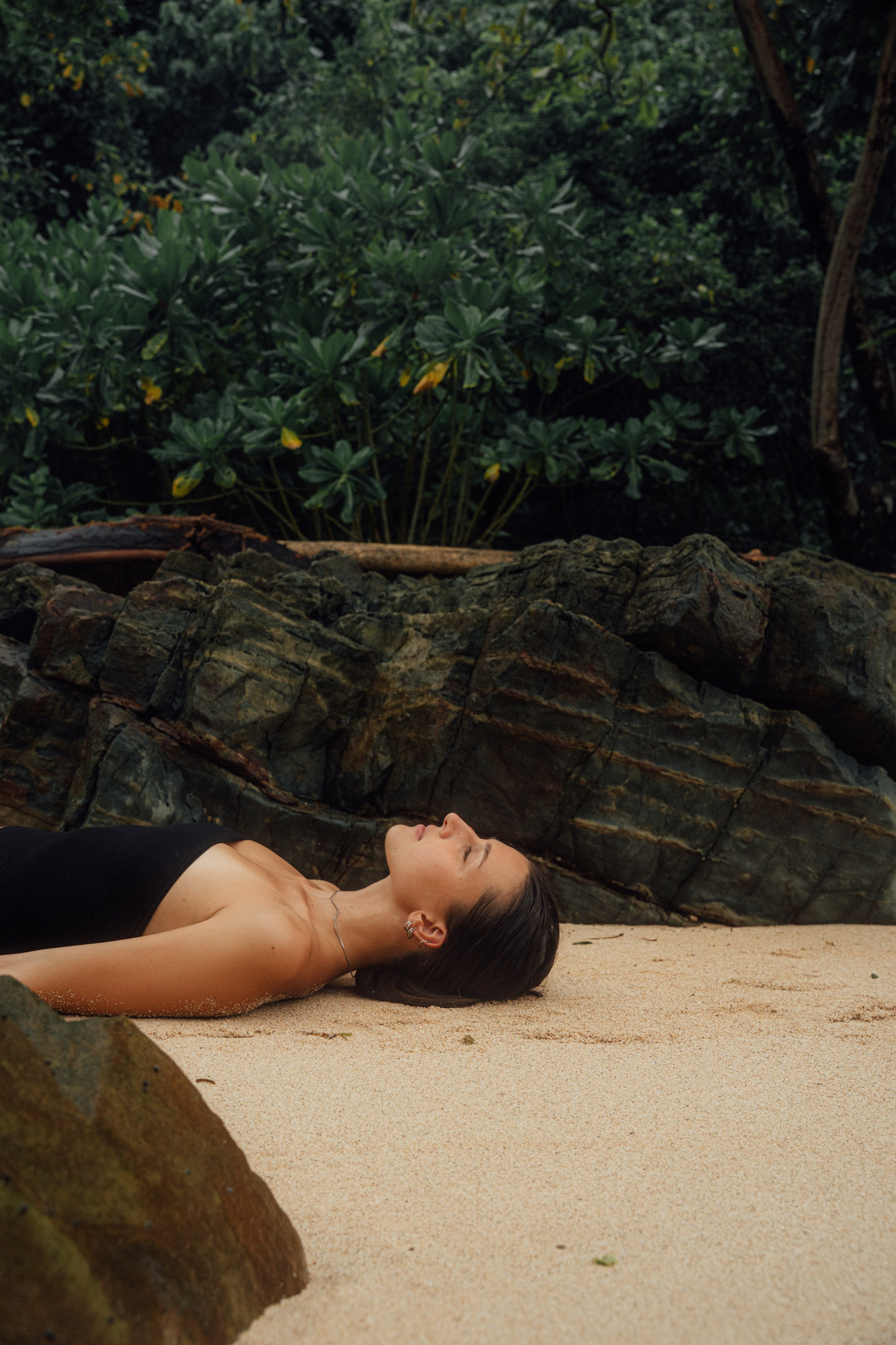 A woman lying on her back on a sandy ground with her eyes closed, surrounded by rocks and green foliage.